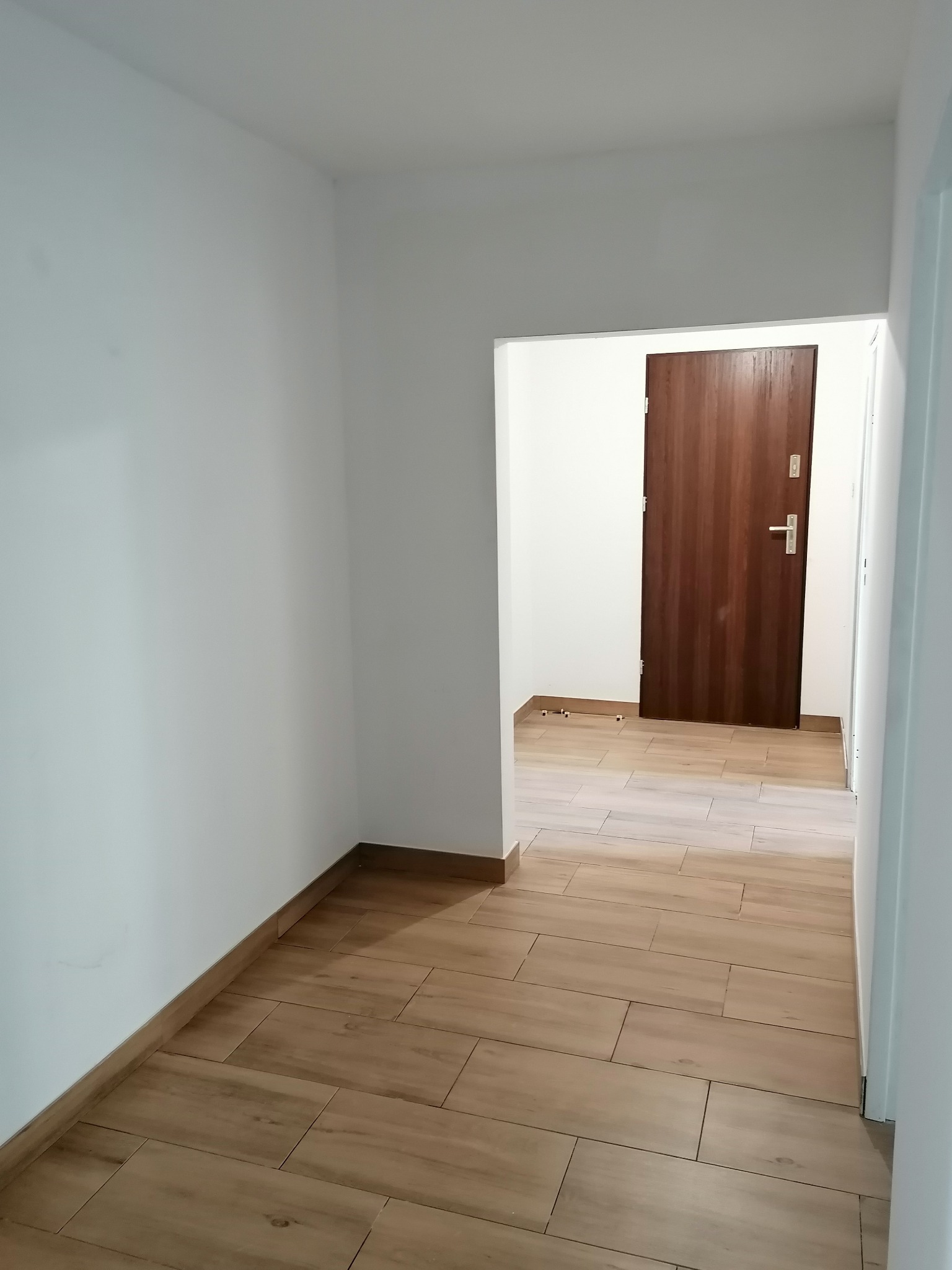 Wykończony korytarz w Gliwicach z drewnopodobną podłogą z płytek i brązowymi drzwiami wejściowymi. Minimalistyczny design, białe ściany.