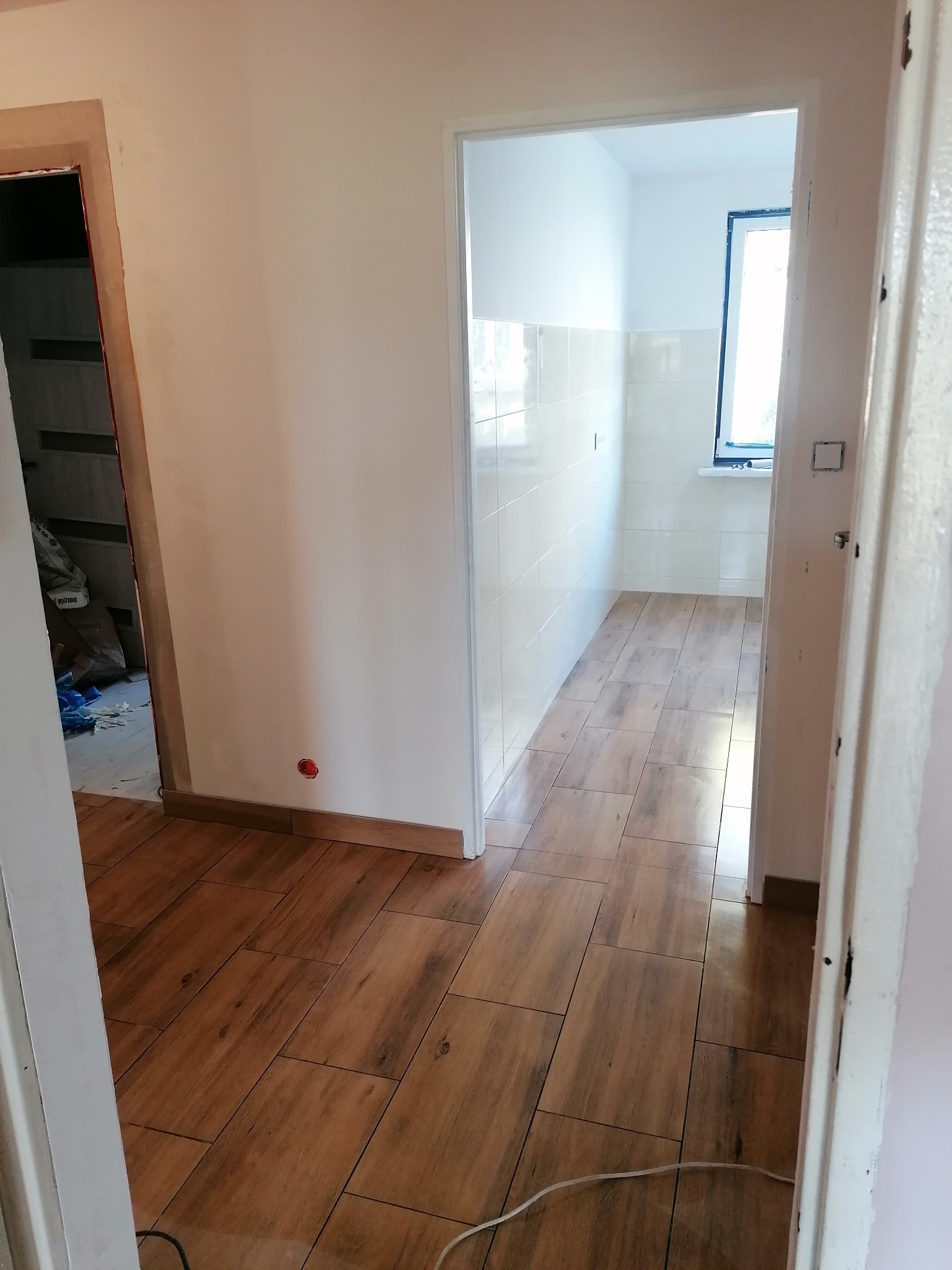 Wykończony korytarz z drewnopodobną podłogą i wejściem do wykafelkowanej łazienki z oknem. Widoczne detale wykończeniowe.