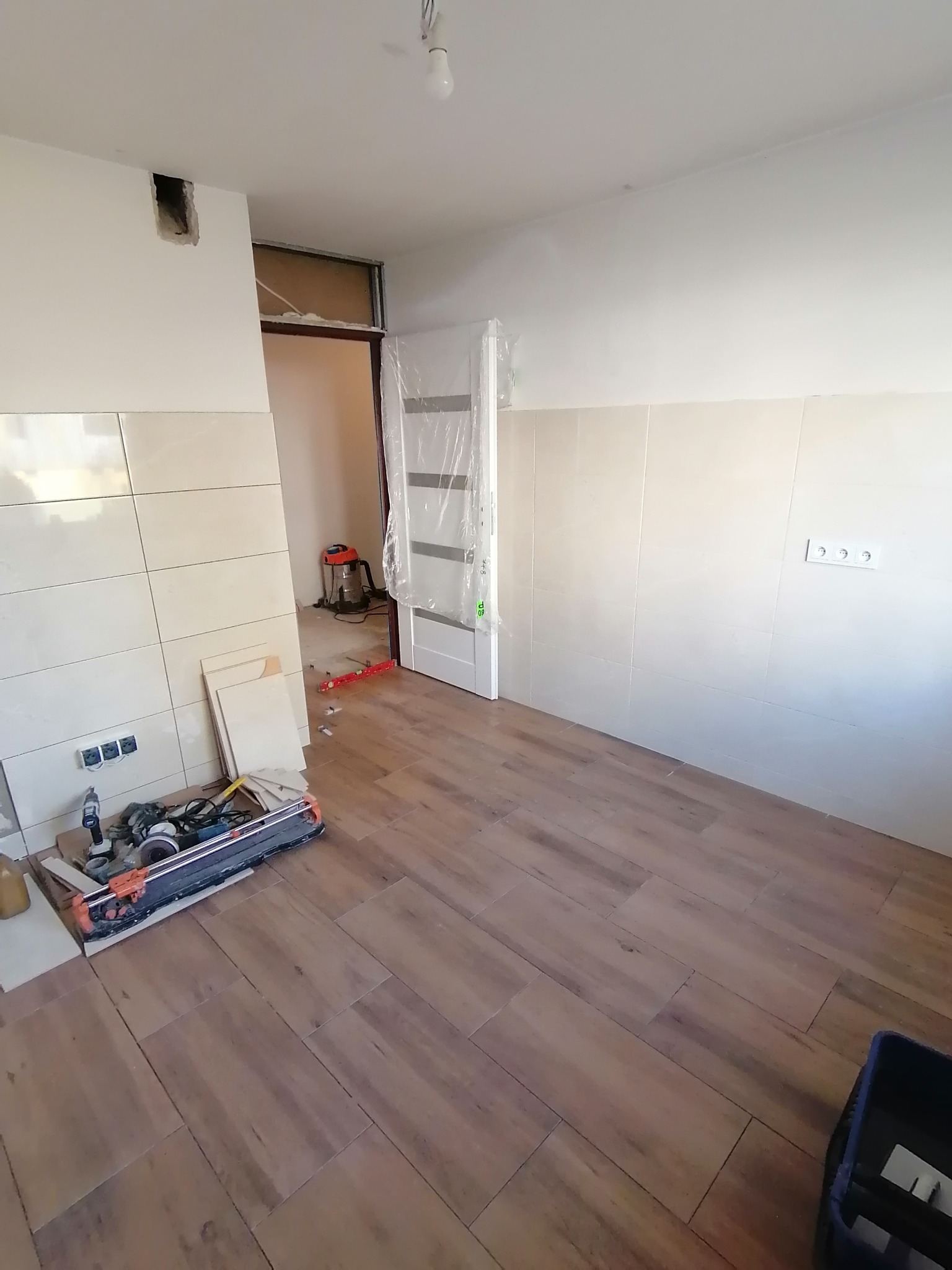 Wnętrze w trakcie układania płytek podłogowych i ściennych. Widoczny sprzęt glazurniczy, fragmenty płytek i gniazdka elektryczne. Częściowo ukończona ściana.