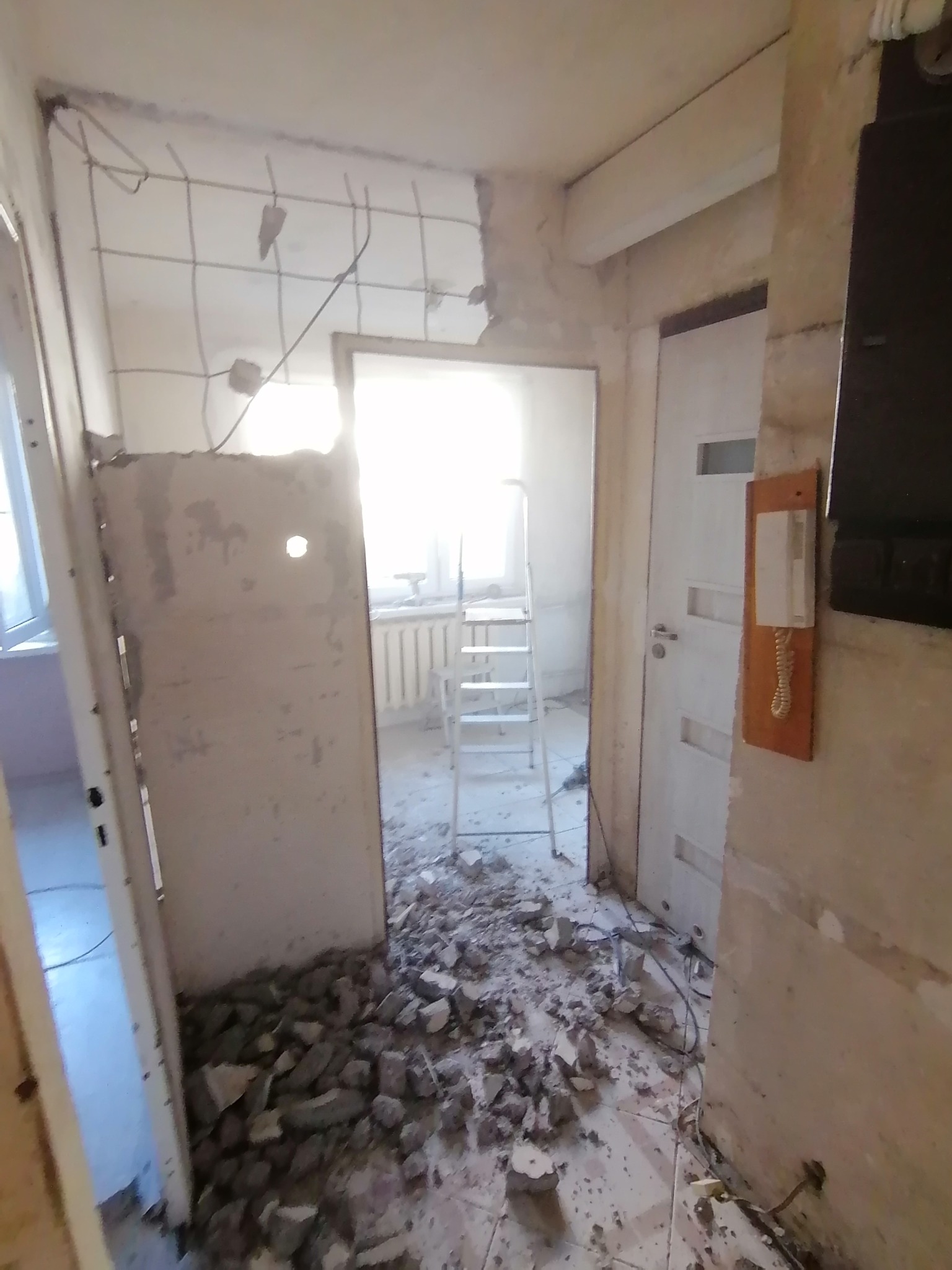 Demontaż ściany działowej: gruz na podłodze, odsłonięte przewody, widok na pokój z drabiną. Etap przygotowawczy do remontu mieszkania.
