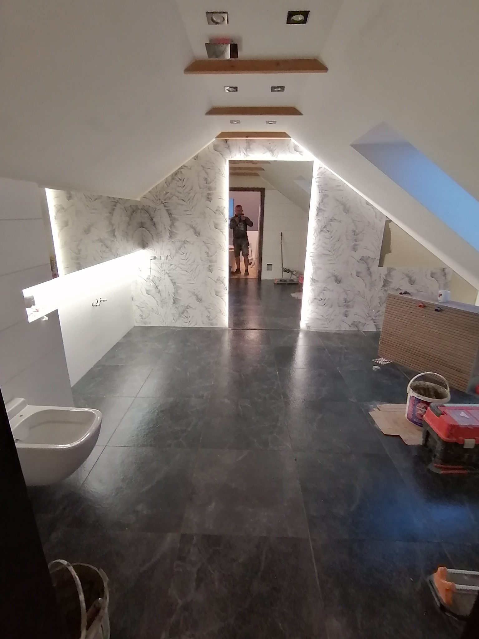 Wnętrze łazienki na poddaszu z szarą marmurkową podłogą i ścianami w biało-szare wzory. Widoczne narzędzia pracy, co sugeruje etap wykończeniowy.