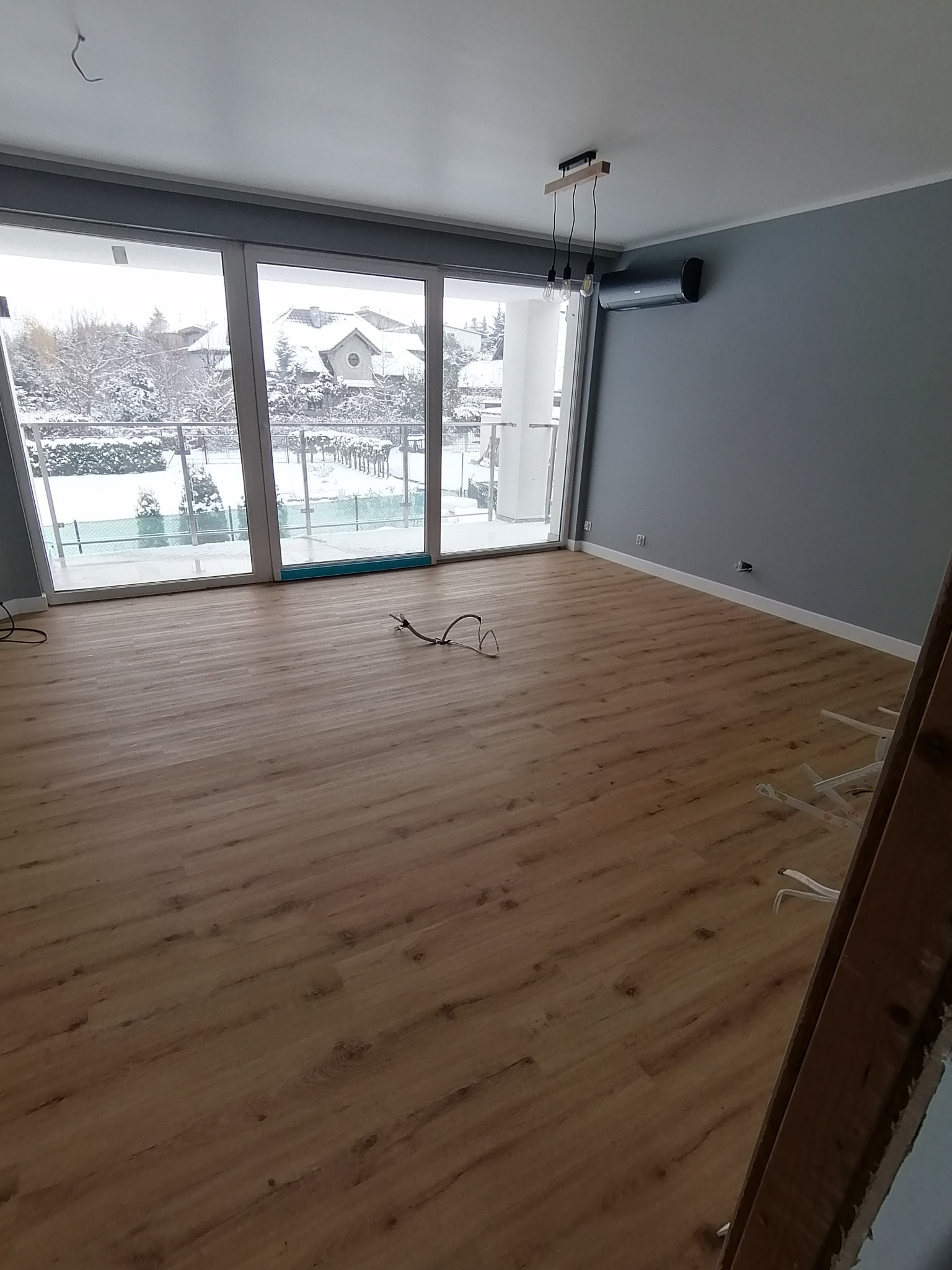 Jasne panele podłogowe w nowym pokoju z dużym oknem balkonowym i szarą ścianą. Widoczne kable i drabina. Za oknem zimowy krajobraz.