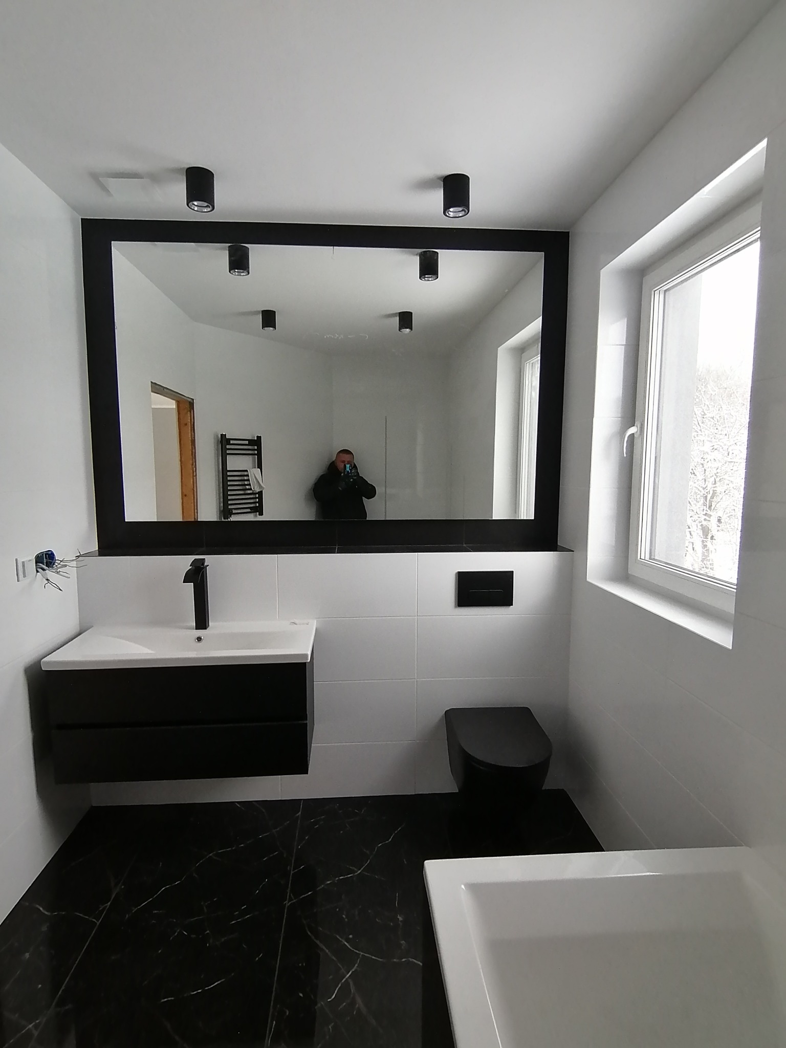 Nowoczesna łazienka z czarną armaturą: umywalka z szafką, toaleta podwieszana, lustro w czarnej ramie. Białe płytki na ścianach i czarna marmurkowa podłoga.