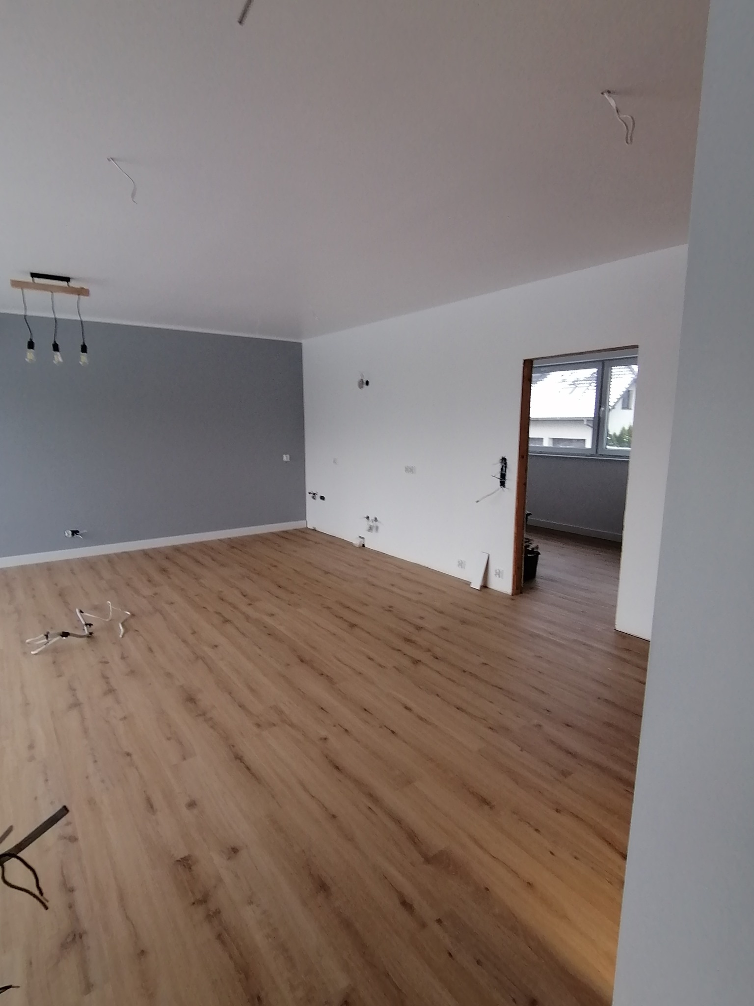 Jasne panele podłogowe w przestronnym pokoju po remoncie, szara ściana z lampą. Widoczne przewody elektryczne i gniazdka, przygotowane do wykończenia. Okno w tle.