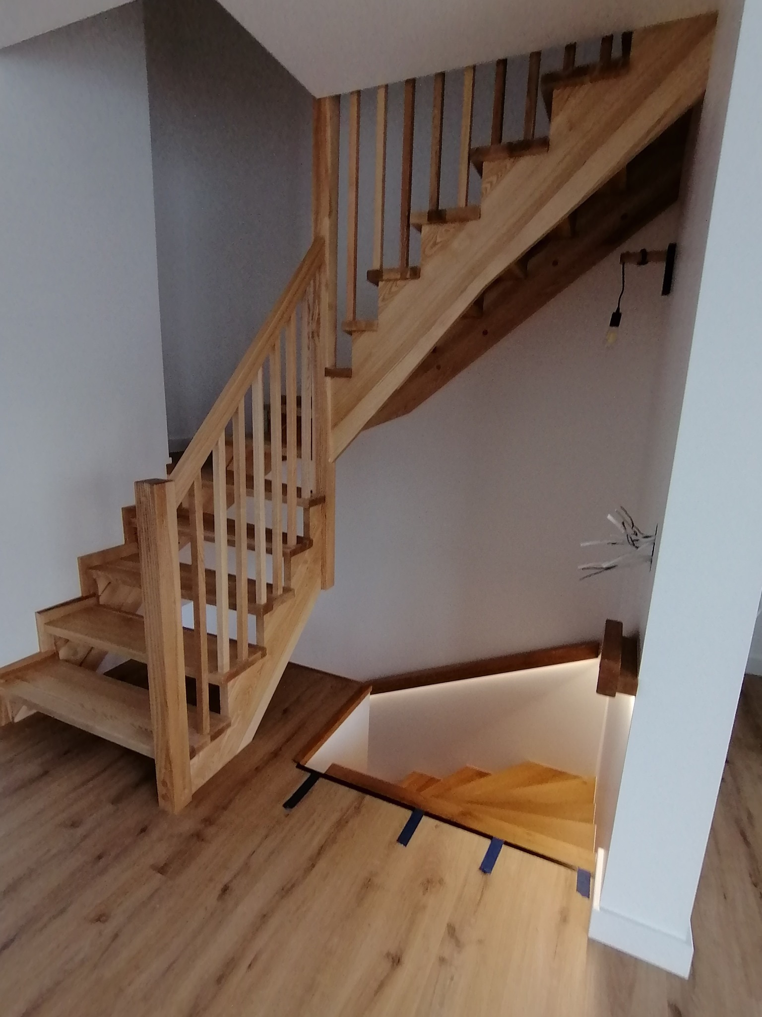 Drewniane schody z podświetleniem LED i barierkami z pionowymi szczeblami, na parkiecie. Widoczne kable elektryczne i lampa na ścianie.