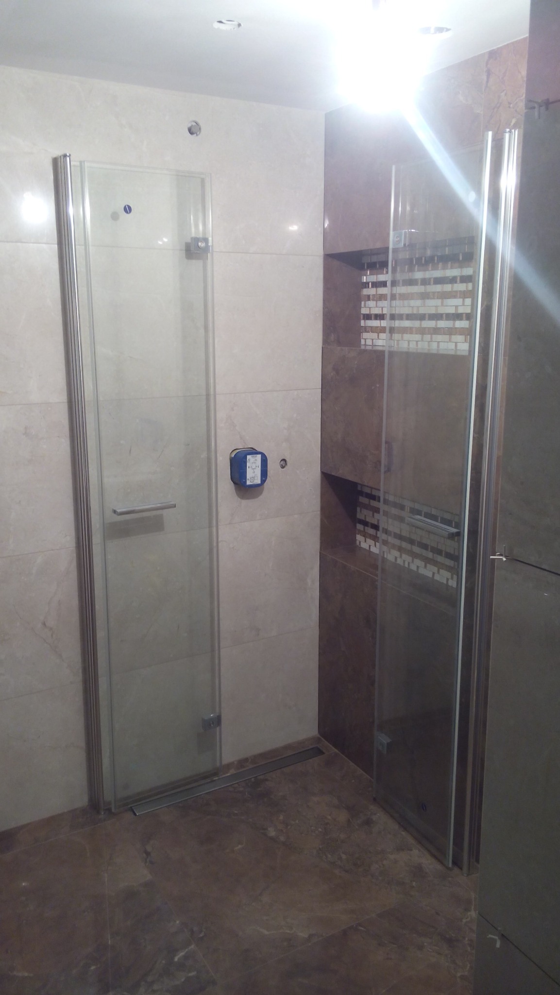 Beżowo-brązowa łazienka z kabiną prysznicową ze szklanymi drzwiami i odpływem liniowym w podłodze, uzupełniona mozaiką na ścianie wnęki.