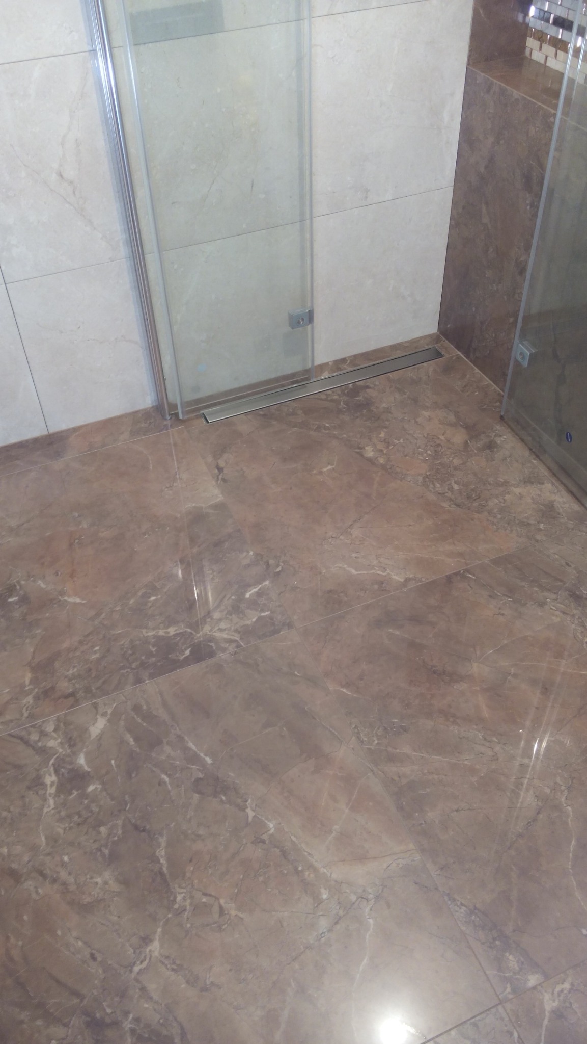 Ukończona łazienka z brązowymi płytkami imitującymi marmur na podłodze i ścianach. Kabina prysznicowa ze szklanymi drzwiami i odpływem liniowym.