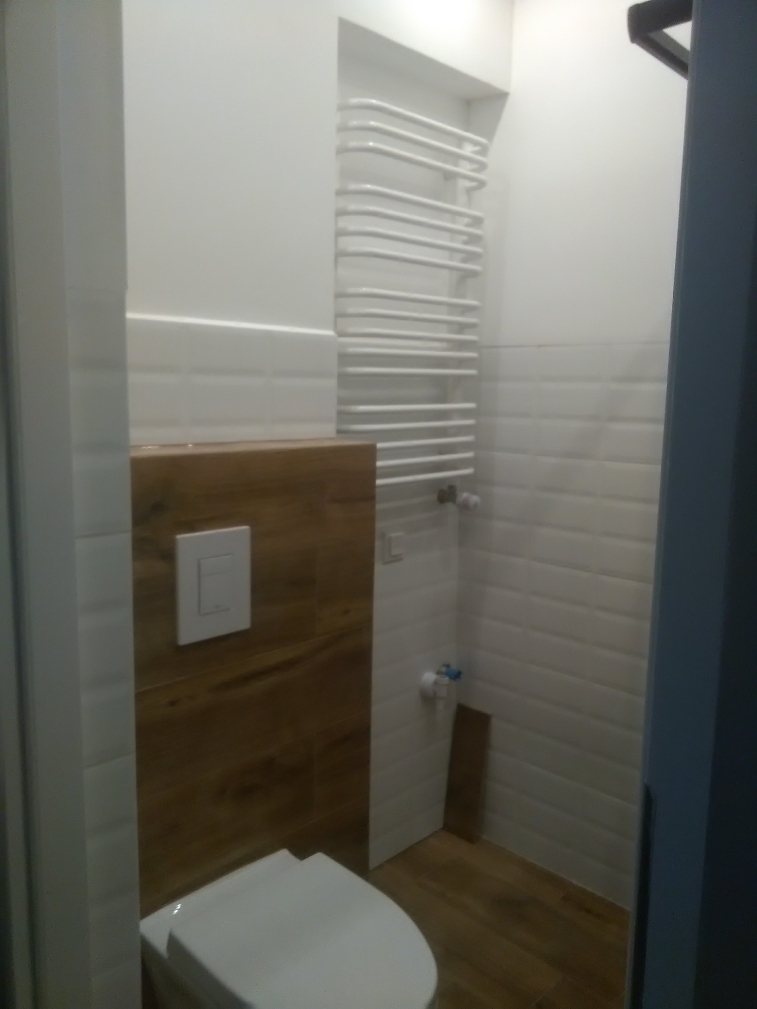 Wykończona łazienka z drewnianym akcentem przy toalecie, białe płytki na ścianach i podłodze, grzejnik drabinkowy. Nowoczesne wnętrze.