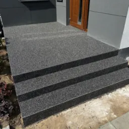 Zewnętrzne schody z posadzką żywiczną w kolorze grafitowym, prowadzące do drzwi wejściowych domu z szarą elewacją. Widok na trzy stopnie i fragment otoczenia.