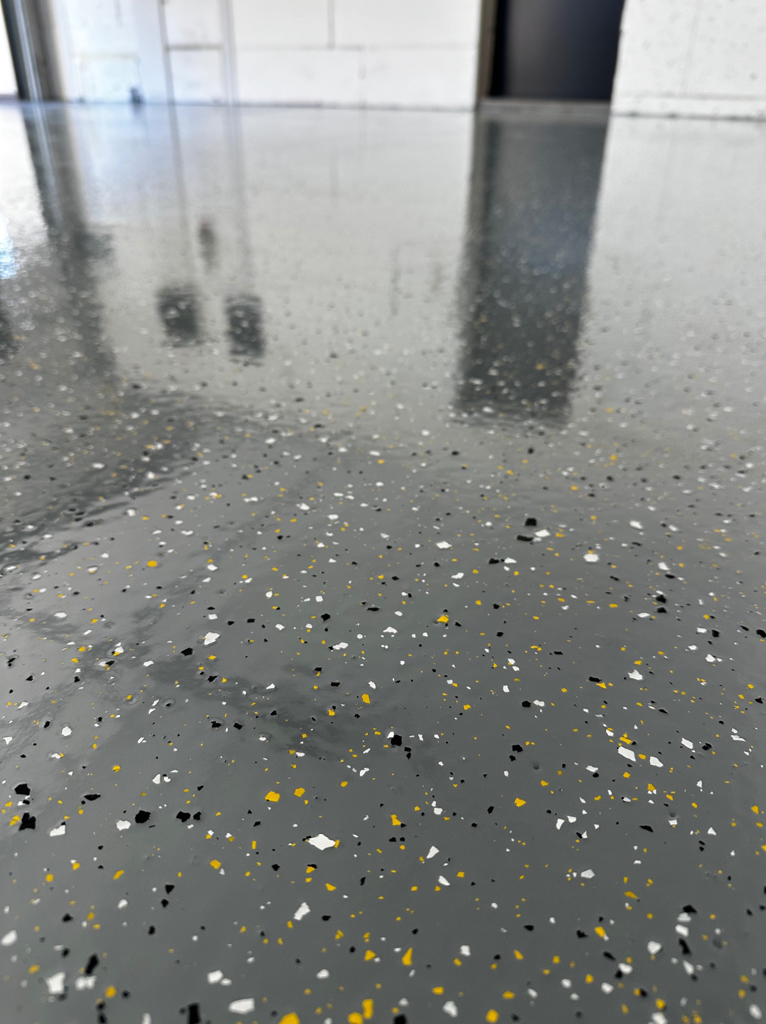 Błyszcząca, szara posadzka z żywicy epoksydowej, z kolorowymi płatkami (białe, czarne, żółte). Gładka powierzchnia odbija światło, widoczne odbicie drzwi i ściany.