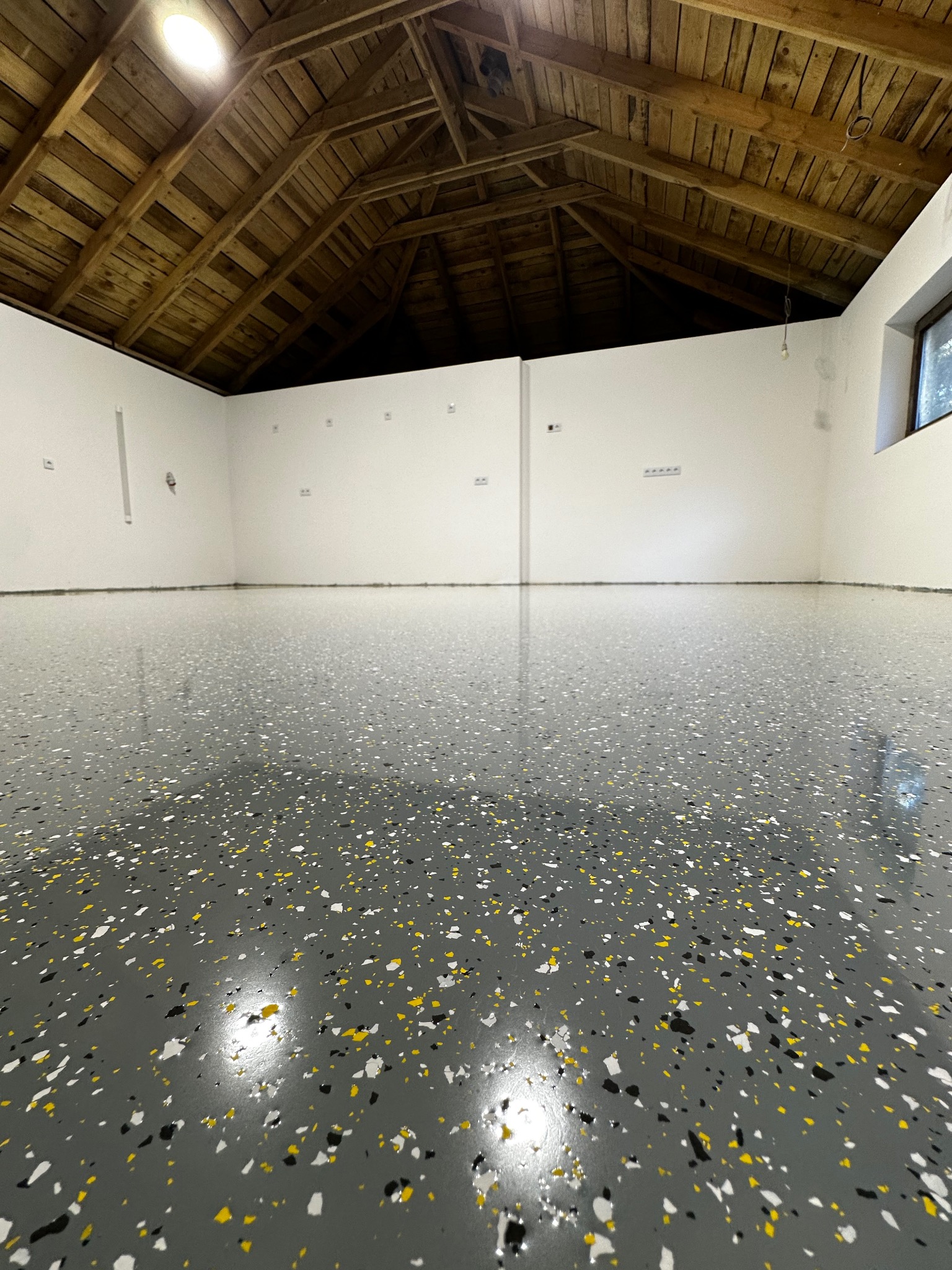 Szara, błyszcząca podłoga z żywicy z kolorowymi płatkami w przestronnym, jasnym pomieszczeniu z drewnianym sufitem i oknem po prawej stronie.