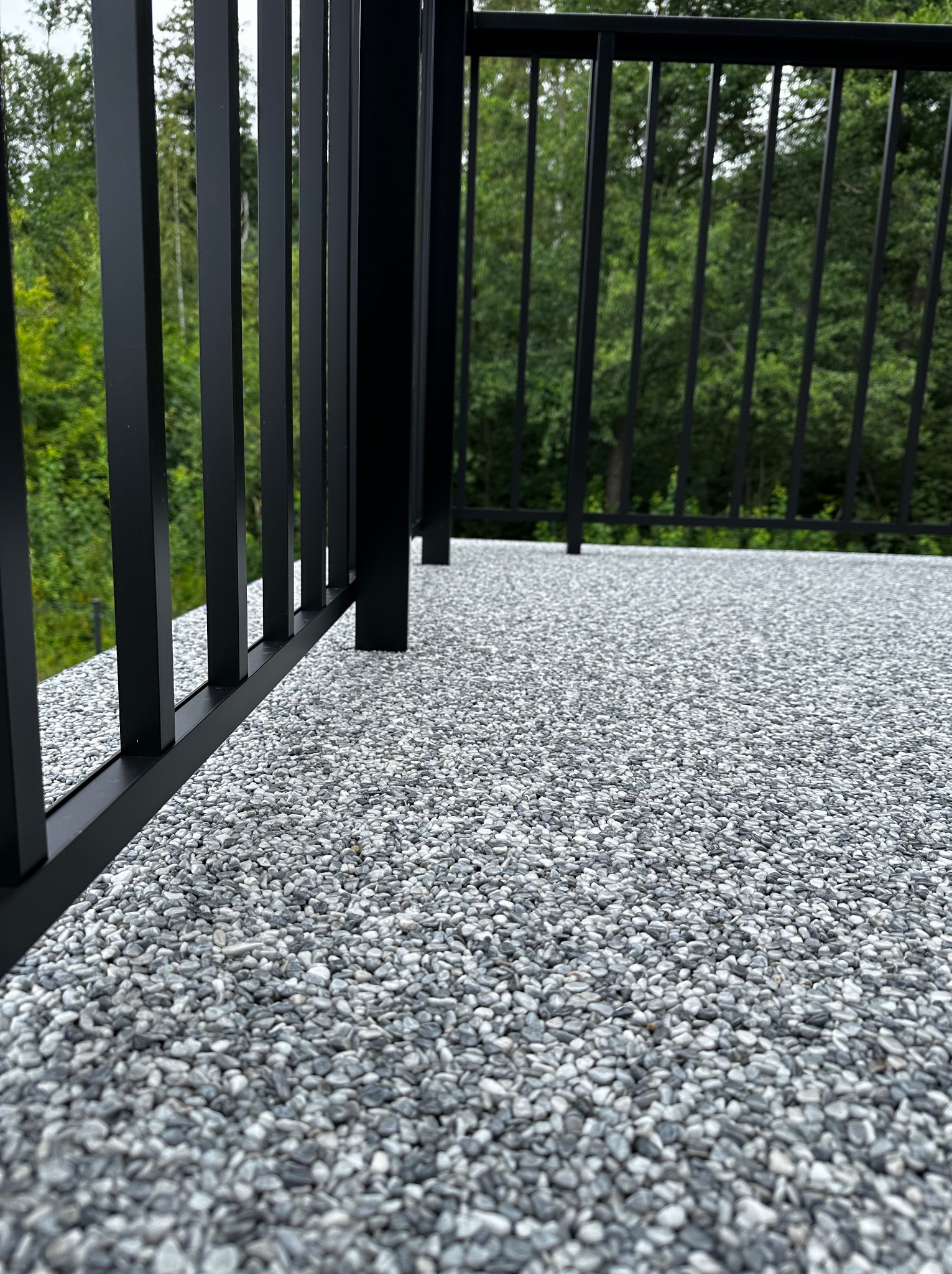 Szara posadzka żywiczna na balkonie z czarną balustradą, widok z bliska na strukturę i zieleń drzew w tle. Realizacja z użyciem kamyczków.