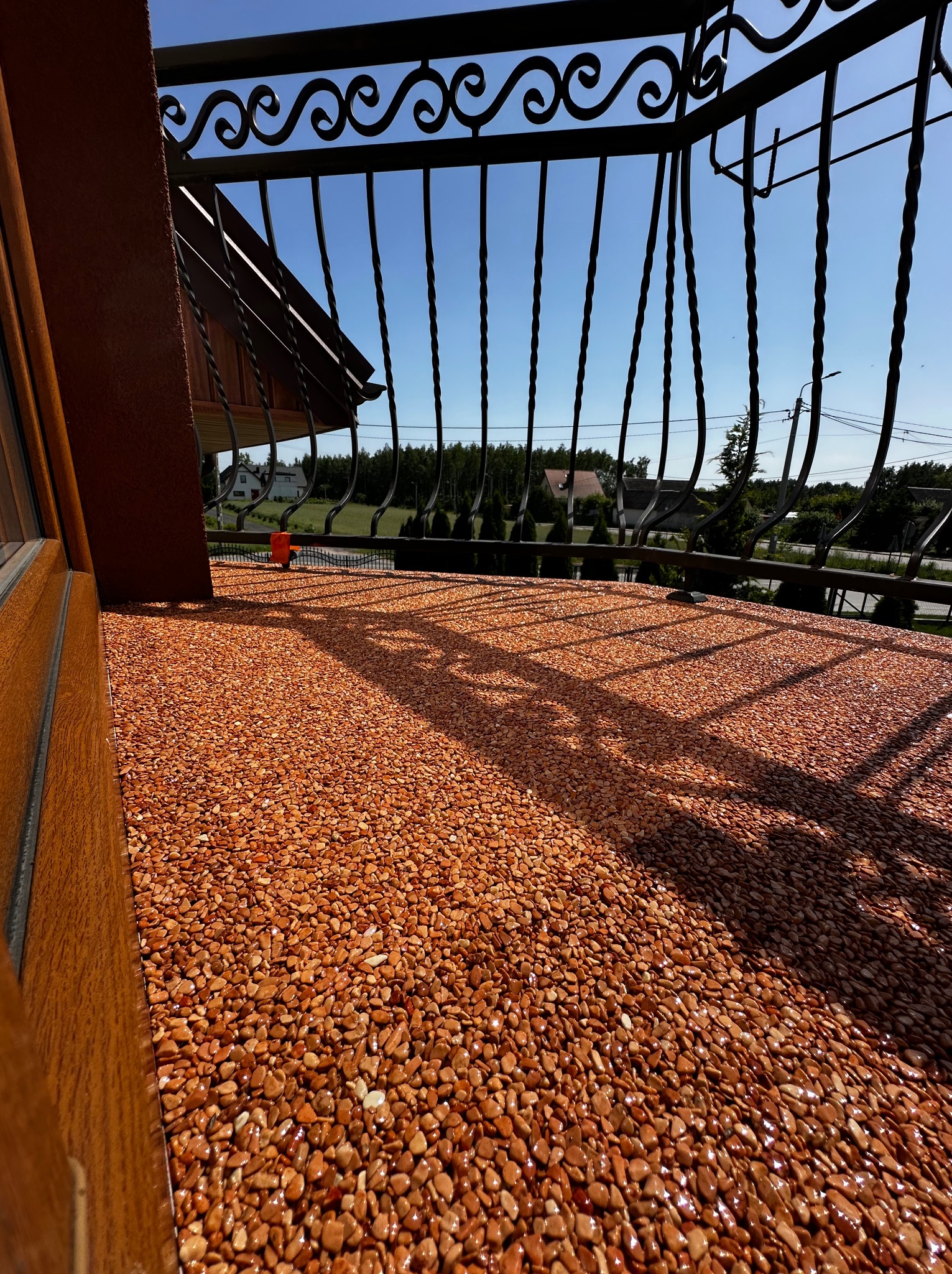Balkon z pomarańczową, kamyczkową nawierzchnią żywiczną w słoneczny dzień. W tle balustrada z kutego żelaza i zieleń.