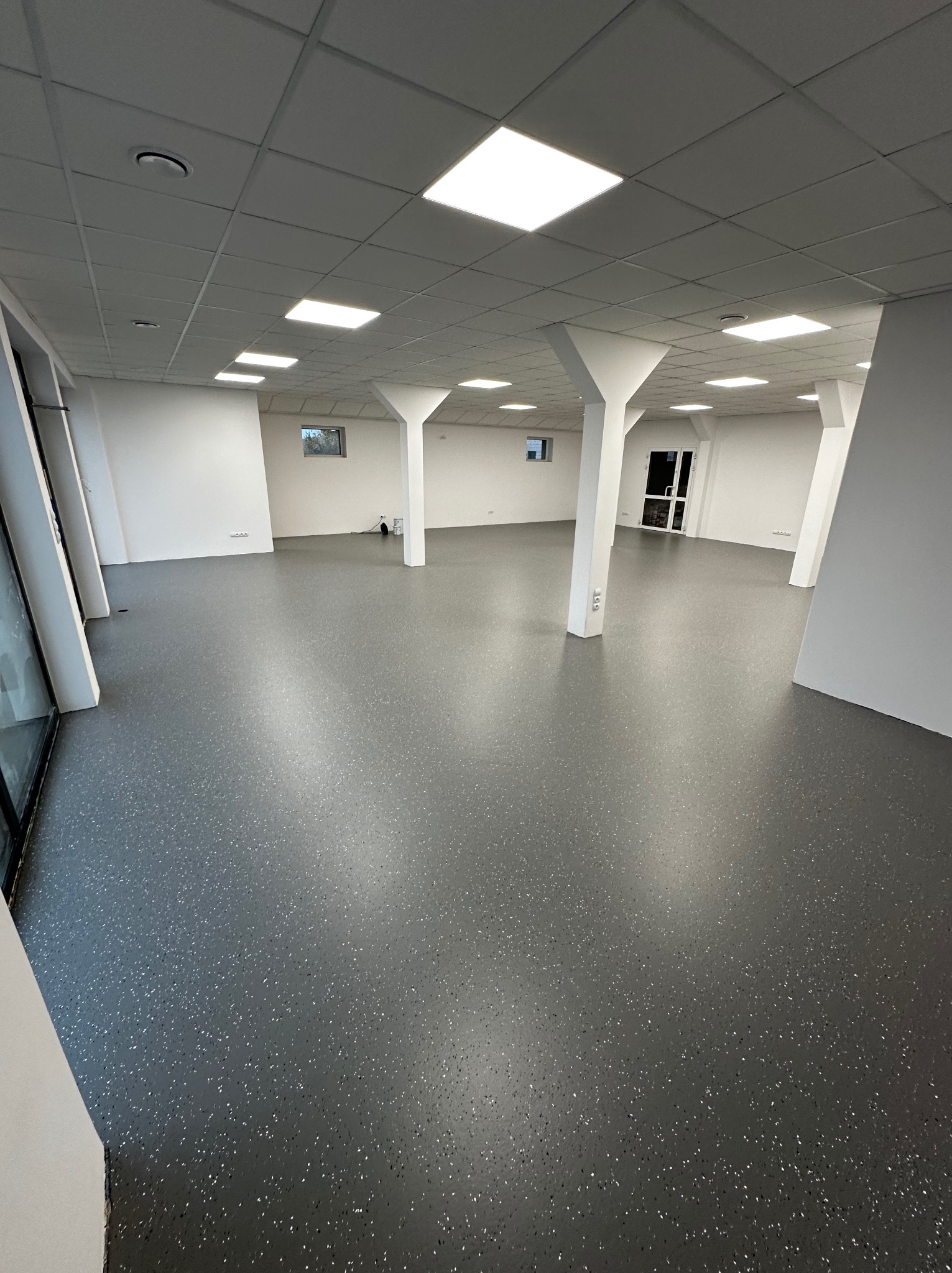 Szara posadzka z drobnymi plamkami w przestronnym, jasnym wnętrzu z białymi kolumnami i oknami. Widok na świeżo wykończoną podłogę.