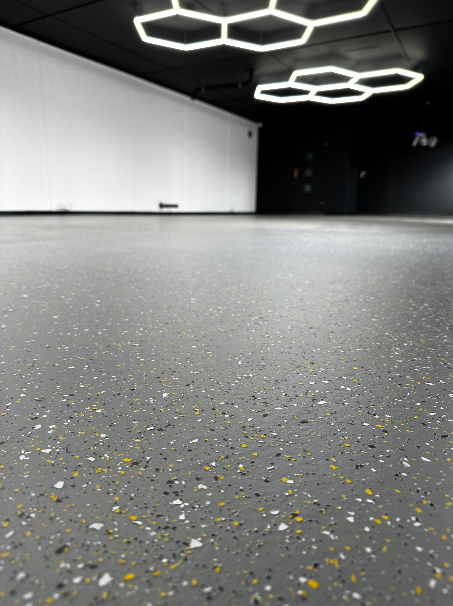 Szara posadzka z kolorowymi płatkami, widoczna z niskiego kąta. W tle biała ściana i heksagonalne lampy sufitowe. Nowoczesne wykończenie podłogi, detal.