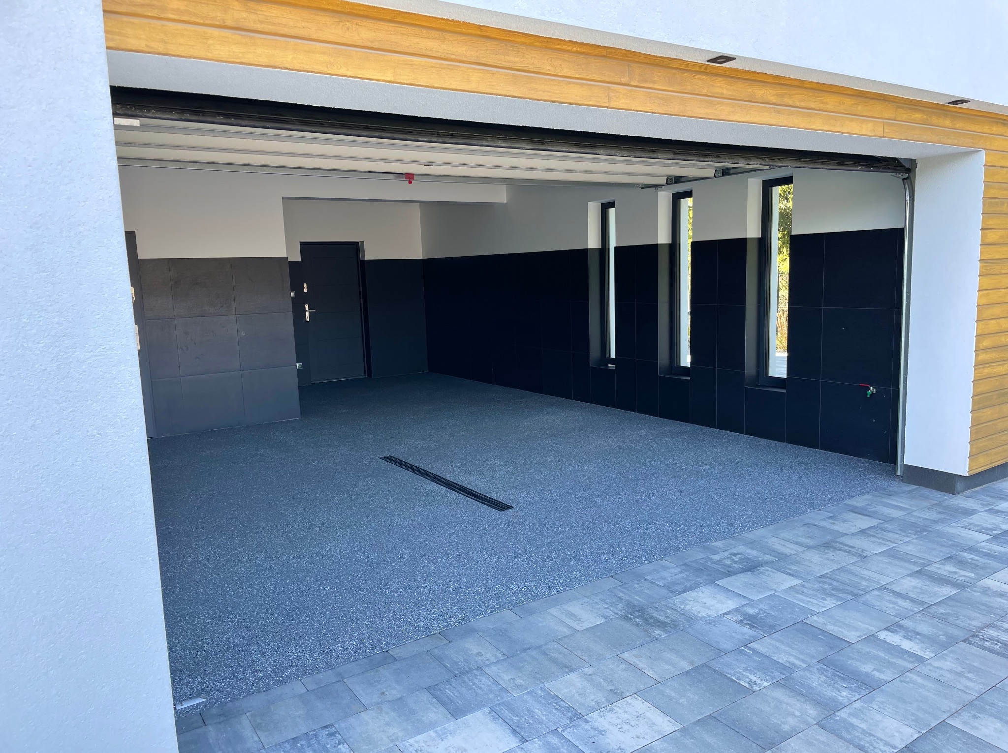Garaż z szarą posadzką żywiczną i odpływem liniowym, widok z zewnątrz. Kontrastowe, czarne ściany i jasne detale architektoniczne.