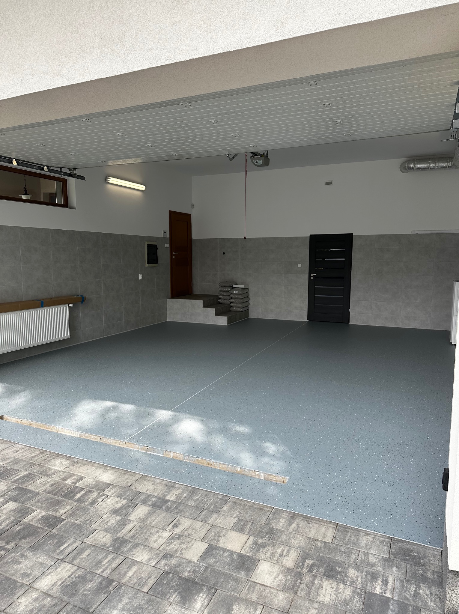Szara posadzka w garażu, wykończona płytkami na zewnątrz. Widoczne płytki ścienne, drzwi i grzejnik. Oświetlenie sufitowe. Nowoczesny design.