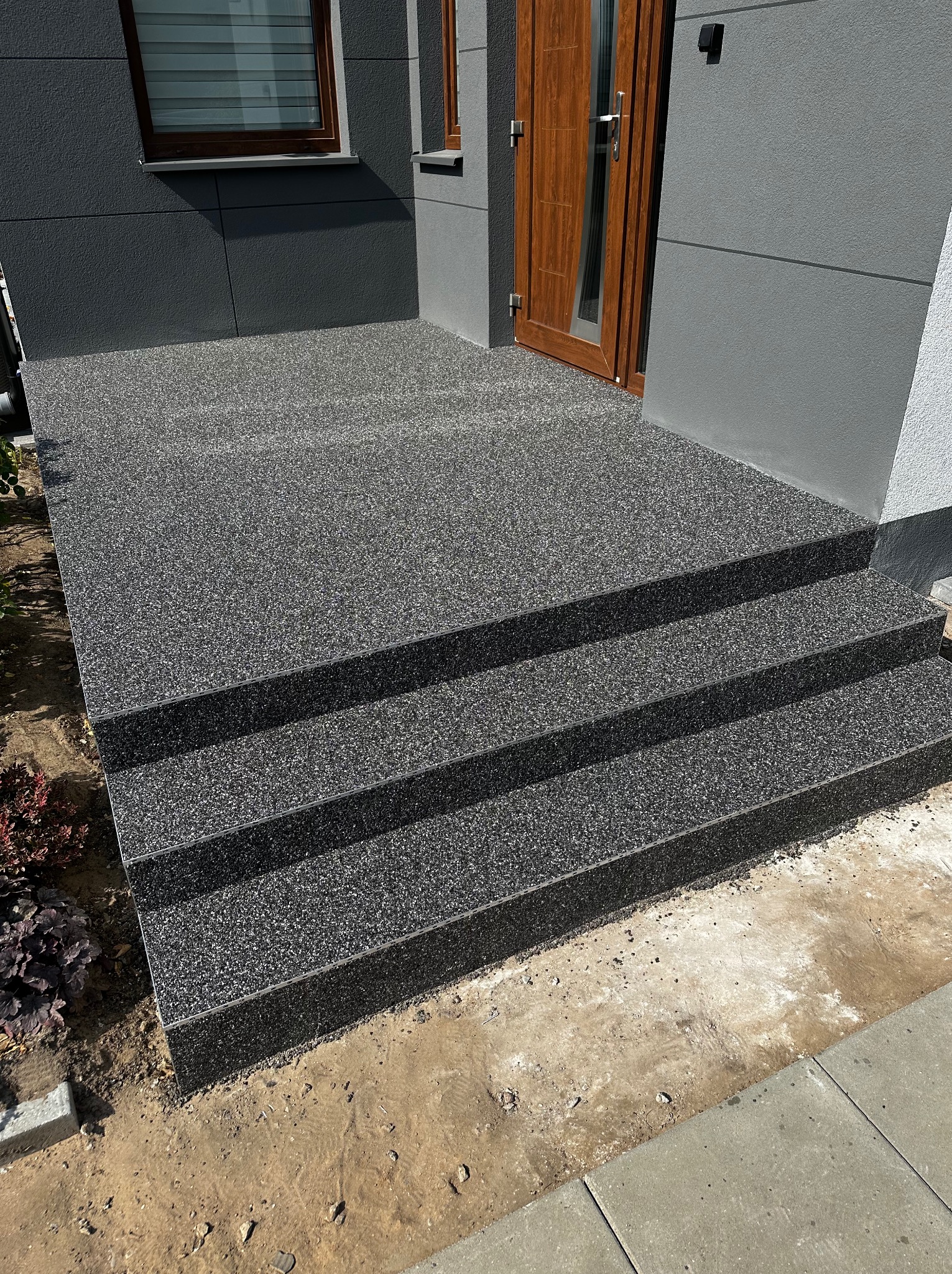 Zewnętrzne schody z posadzką żywiczną w kolorze grafitowym, prowadzące do drzwi wejściowych domu z szarą elewacją. Widok na trzy stopnie i fragment otoczenia.