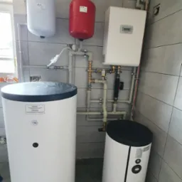 Kompletna instalacja grzewcza w kotłowni: dwa zbiorniki akumulacyjne, ekspansomat czerwony i biały, piec gazowy. Widoczne rury i zawory. Szare płytki na ścianach i podłodze.