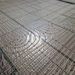 Ułożone rury ogrzewania podłogowego na folii z czerwoną siatką, przymocowane czarnymi uchwytami, gotowe do zalania posadzką w surowym pomieszczeniu.