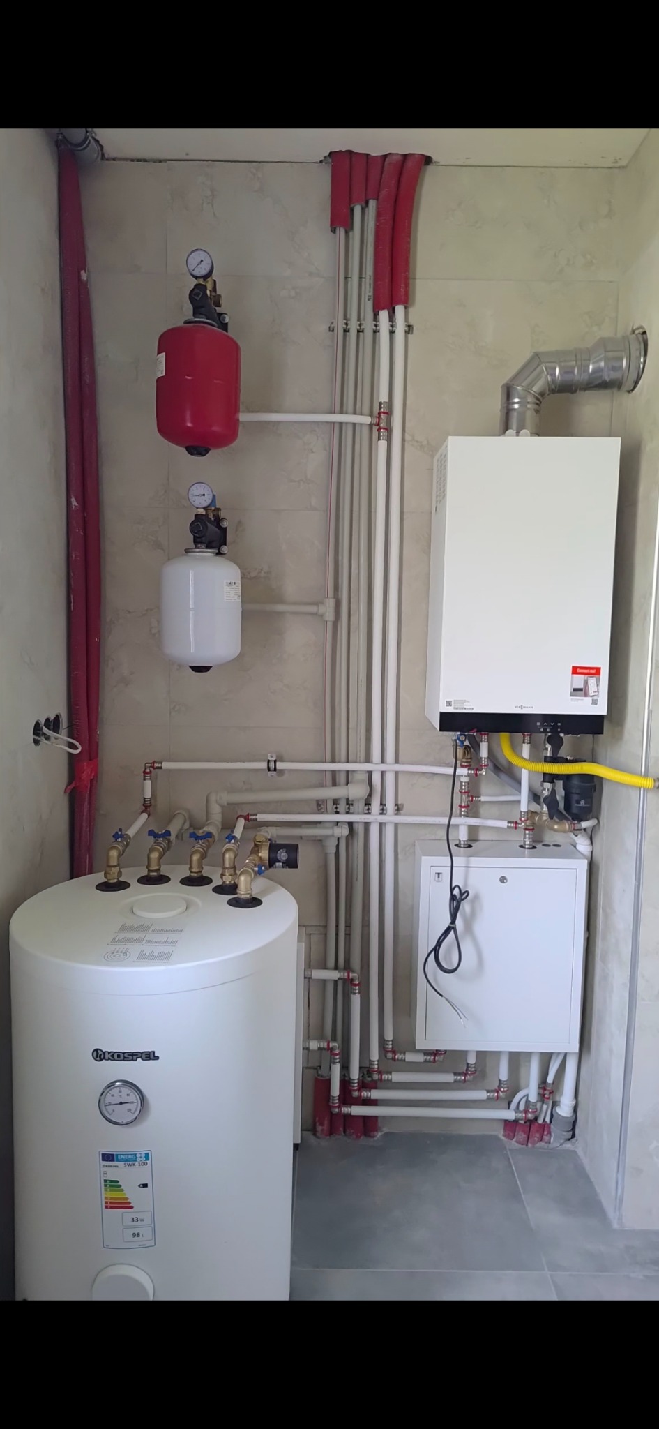 Złożona instalacja grzewcza w kotłowni: biały piec gazowy Viessmann, zbiornik Kospel, czerwone naczynia wzbiorcze, liczne rury i zawory na tle marmurowej ściany.