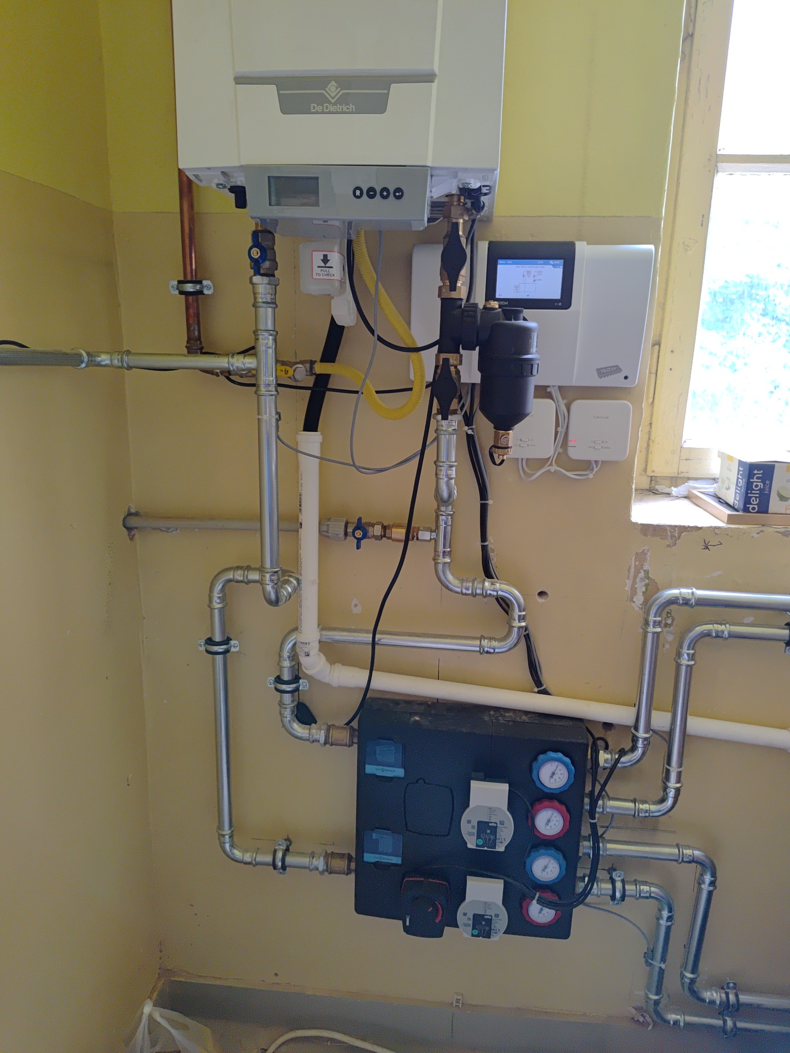 Nowoczesny piec gazowy De Dietrich z podłączeniami, widoczny panel sterowania i liczne rury instalacyjne, zamontowany na żółtej ścianie.