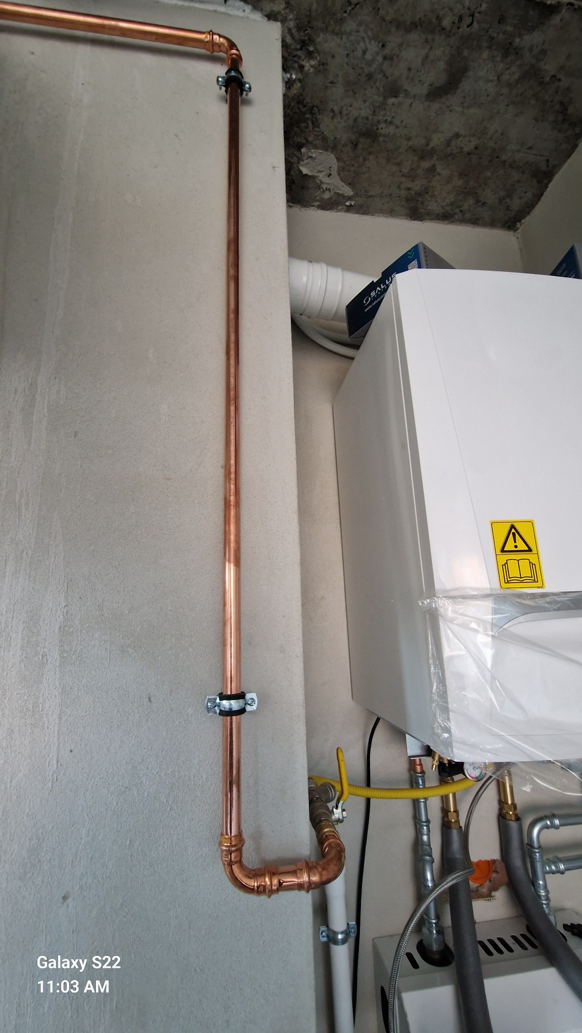 Miedziana instalacja gazowa z rurami przymocowanymi do ściany, obok biały piec gazowy z żółtą naklejką ostrzegawczą. Widoczne szare ściany i sufit.