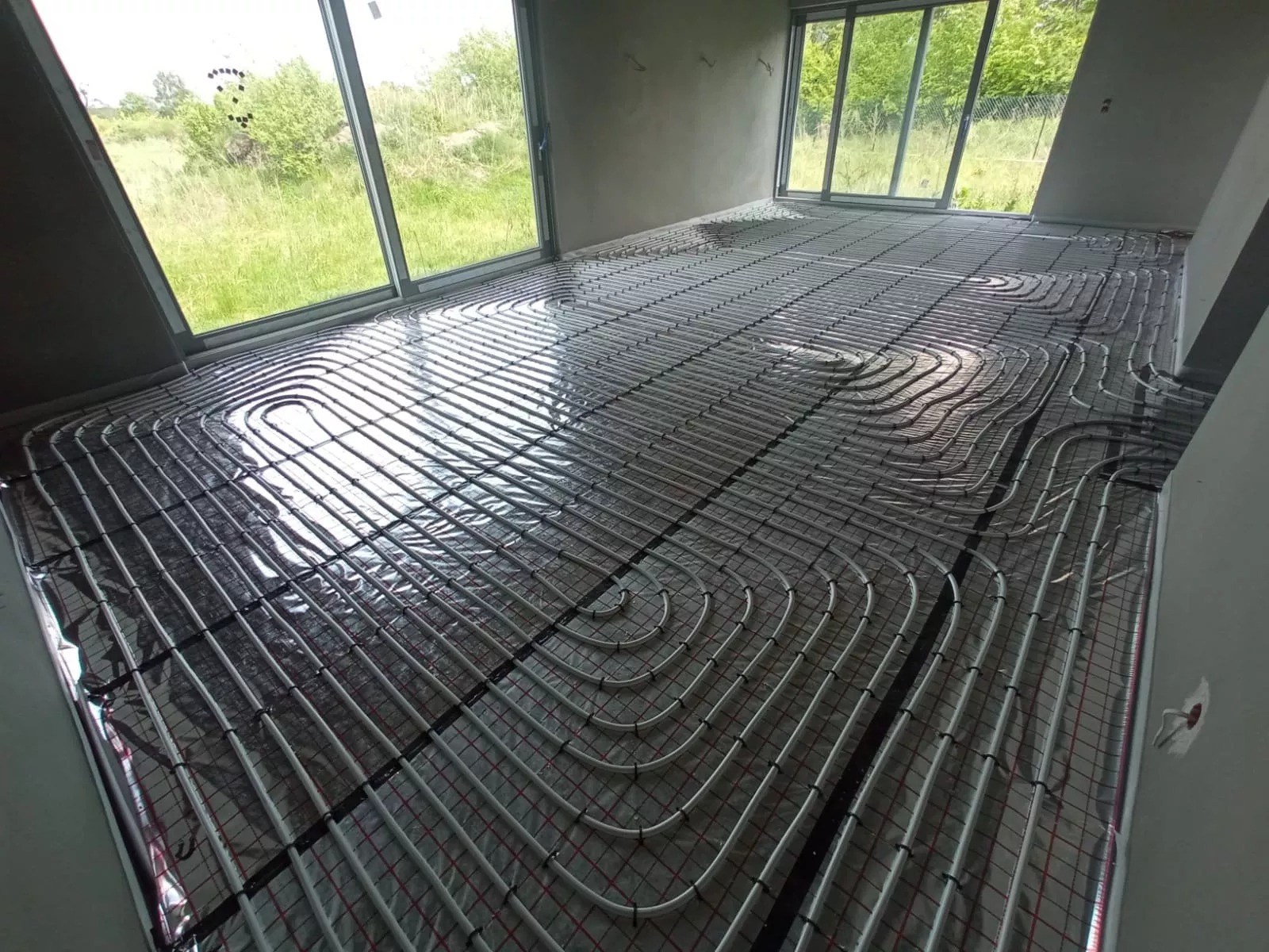 Białe rury ogrzewania podłogowego ułożone na folii i siatce w jasnym pokoju z dużymi oknami. Widok z góry ukazuje spiralny układ rur.