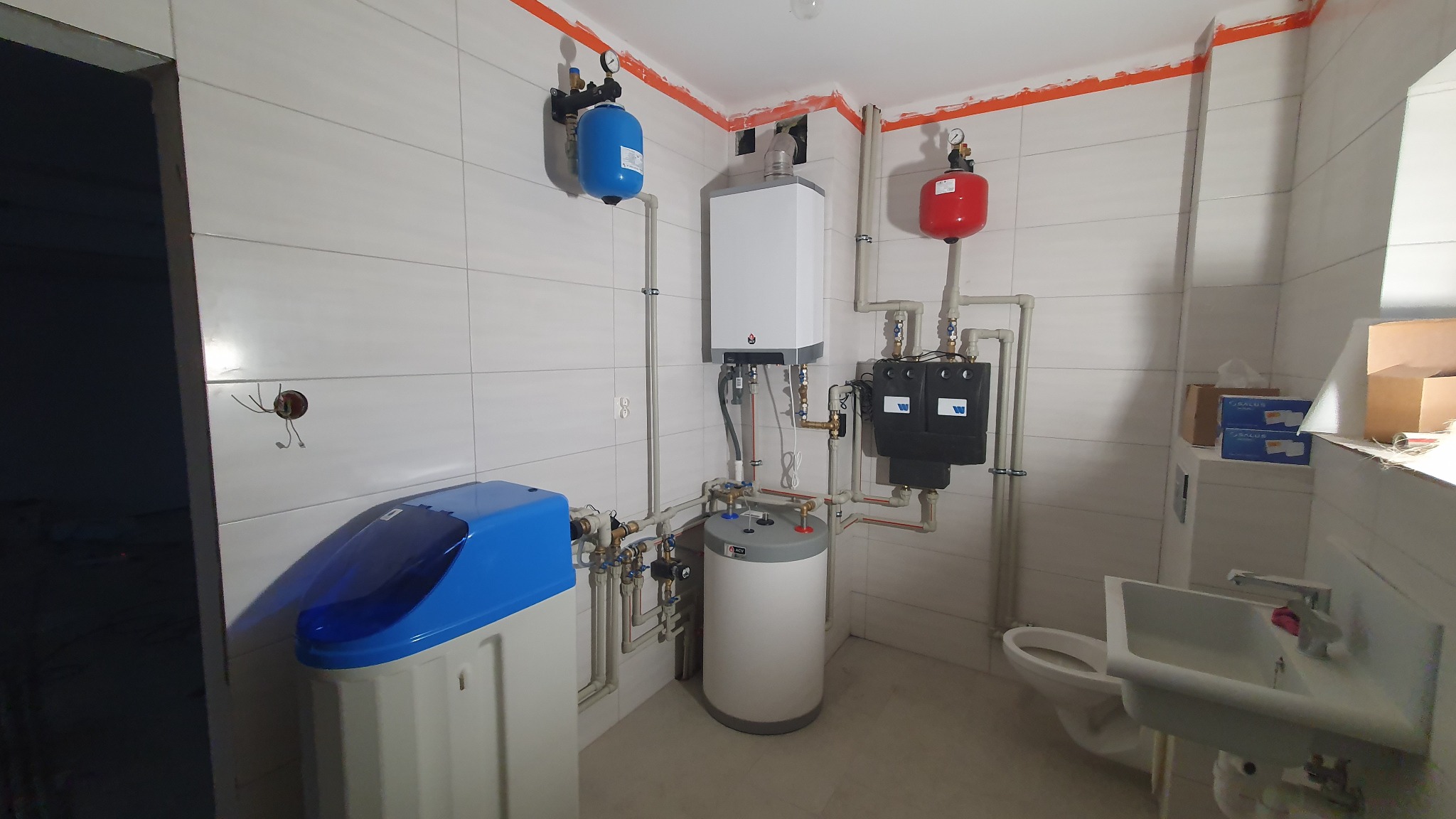 Nowoczesna kotłownia w jasnej, wykafelkowanej łazience z niebieskim zmiękczaczem wody i białym piecem gazowym na ścianie, otoczona rurami i armaturą.