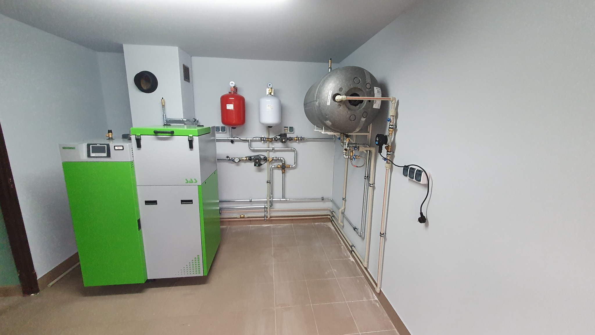 Kompletna instalacja grzewcza z nowoczesnym piecem na pellet, zbiornikiem akumulacyjnym i naczyniami przeponowymi w kotłowni z wykafelkowaną podłogą.