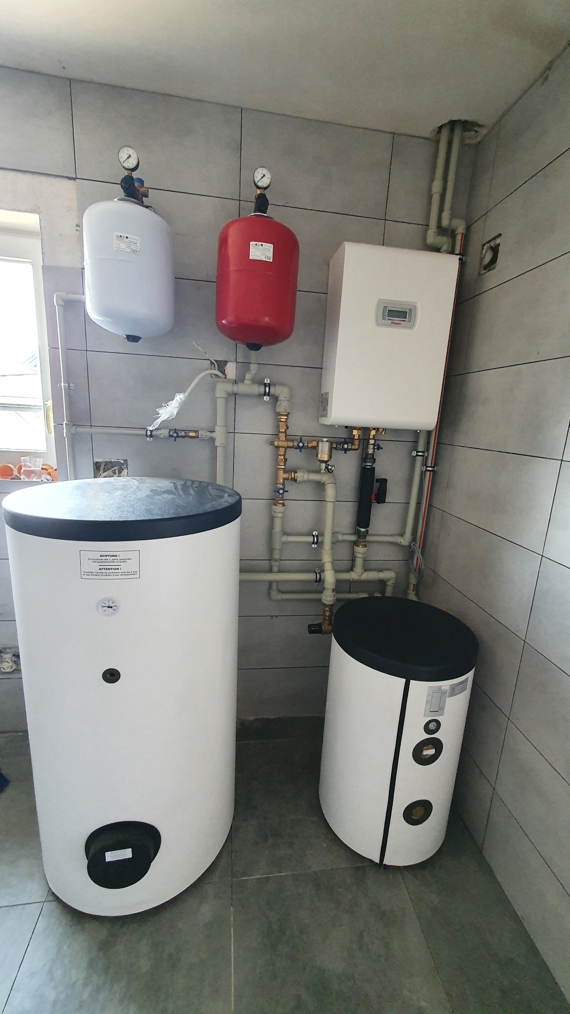 Kompletna instalacja grzewcza w kotłowni: dwa zbiorniki akumulacyjne, ekspansomat czerwony i biały, piec gazowy. Widoczne rury i zawory. Szare płytki na ścianach i podłodze.