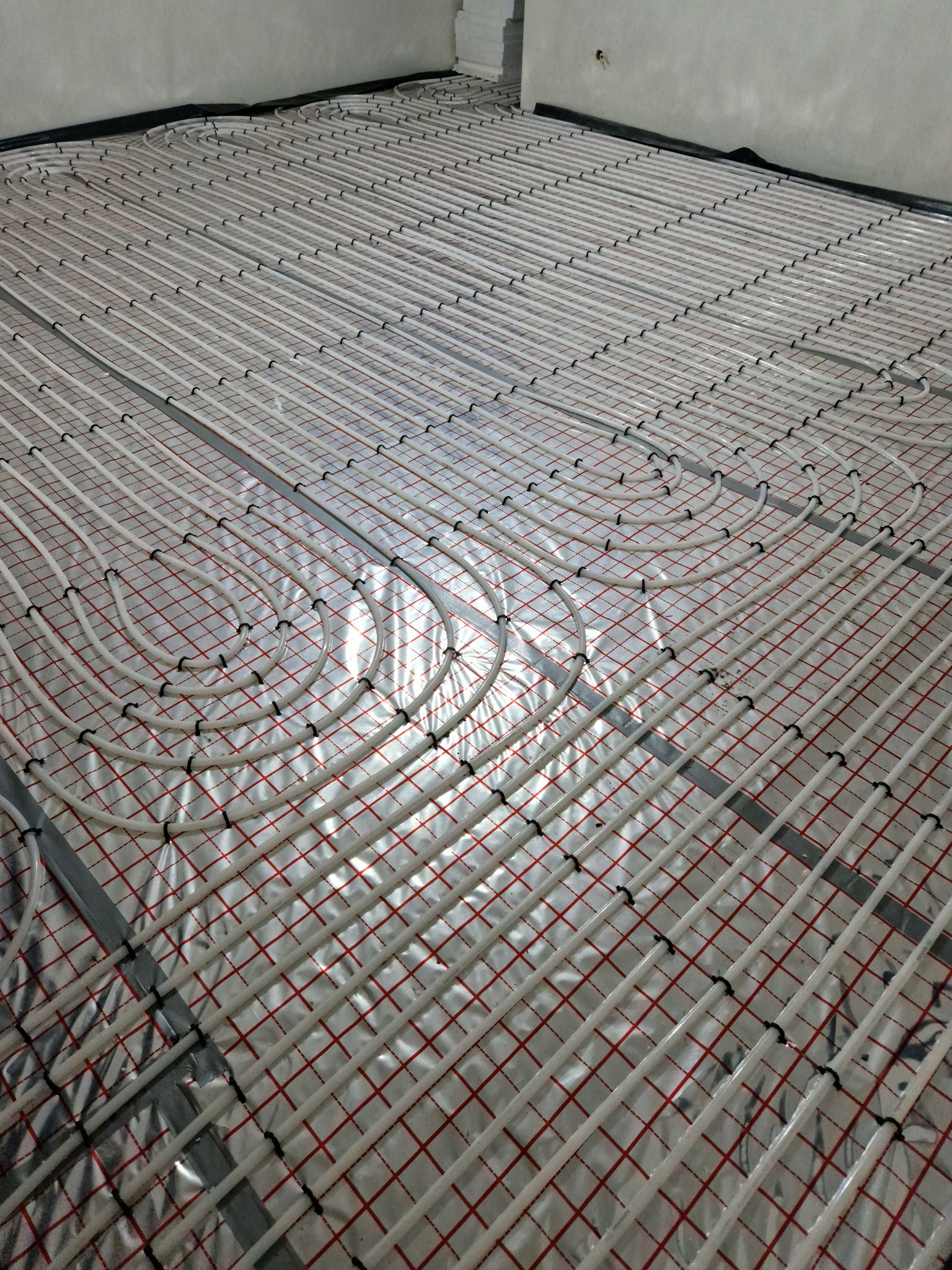 Ułożone rury ogrzewania podłogowego na folii z czerwoną siatką, przymocowane czarnymi uchwytami, gotowe do zalania posadzką w surowym pomieszczeniu.