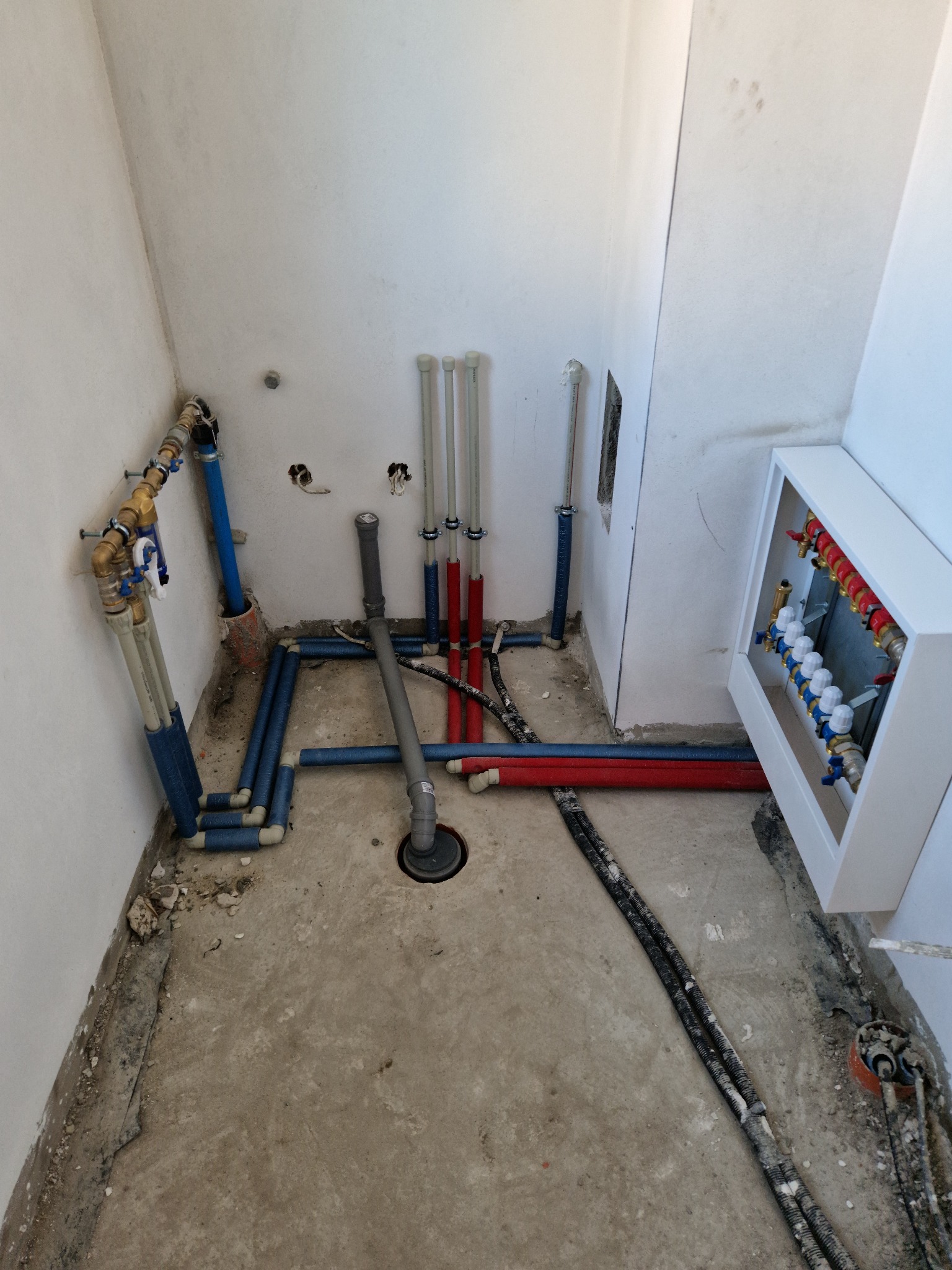 Wnętrze łazienki w trakcie instalacji wodno-kanalizacyjnej z rozdzielaczem, rurami w izolacji i odpływem podłogowym. Widoczne przyłącza i fragment ściany.