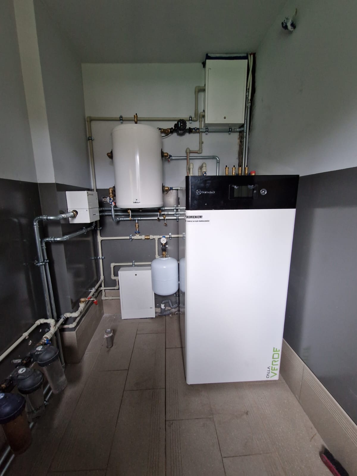 Nowoczesny, biało-czarny piec grzewczy z podłączonymi rurami w pomieszczeniu z szaro-białymi ścianami i brązową podłogą. Widoczne filtry i zbiornik.