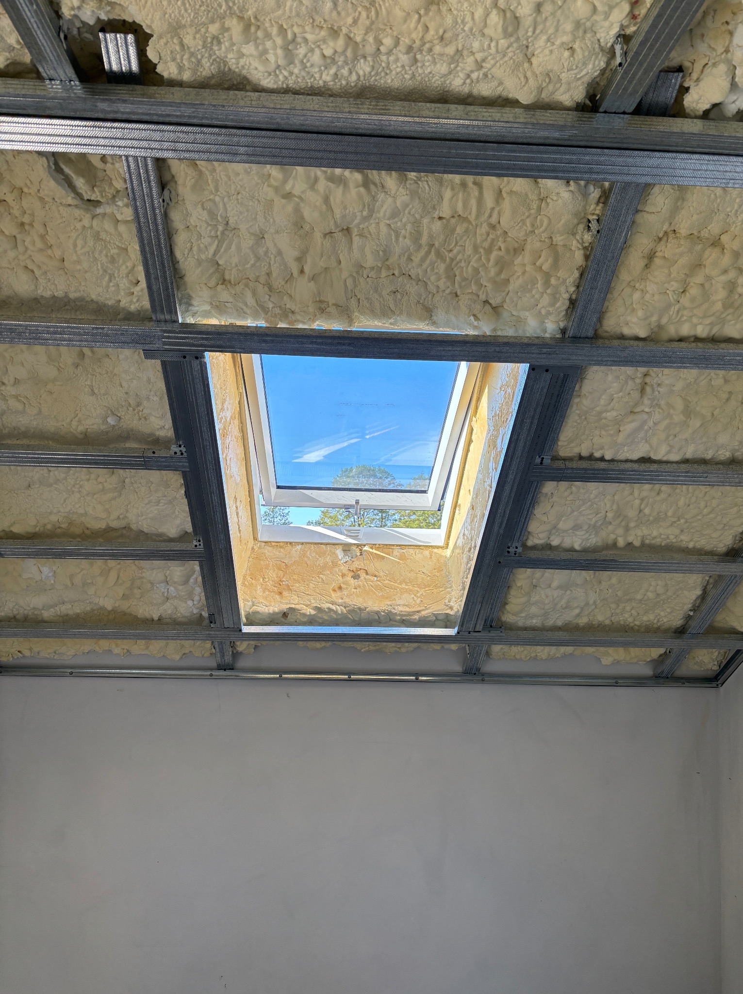 Metalowy stelaż sufitowy z oknem dachowym i pianką izolacyjną, widok z dołu, jasne wnętrze w trakcie prac wykończeniowych.