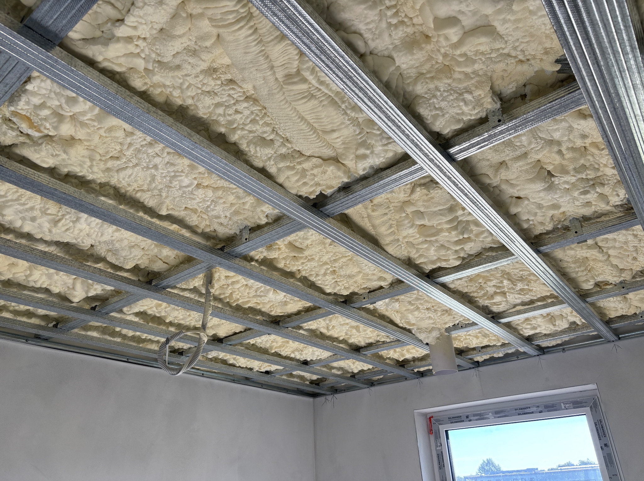 Ocieplenie pianą PUR pod metalowym rusztem sufitu podwieszanego. Widoczne przewody elektryczne i okno. Szara ściana w tle.