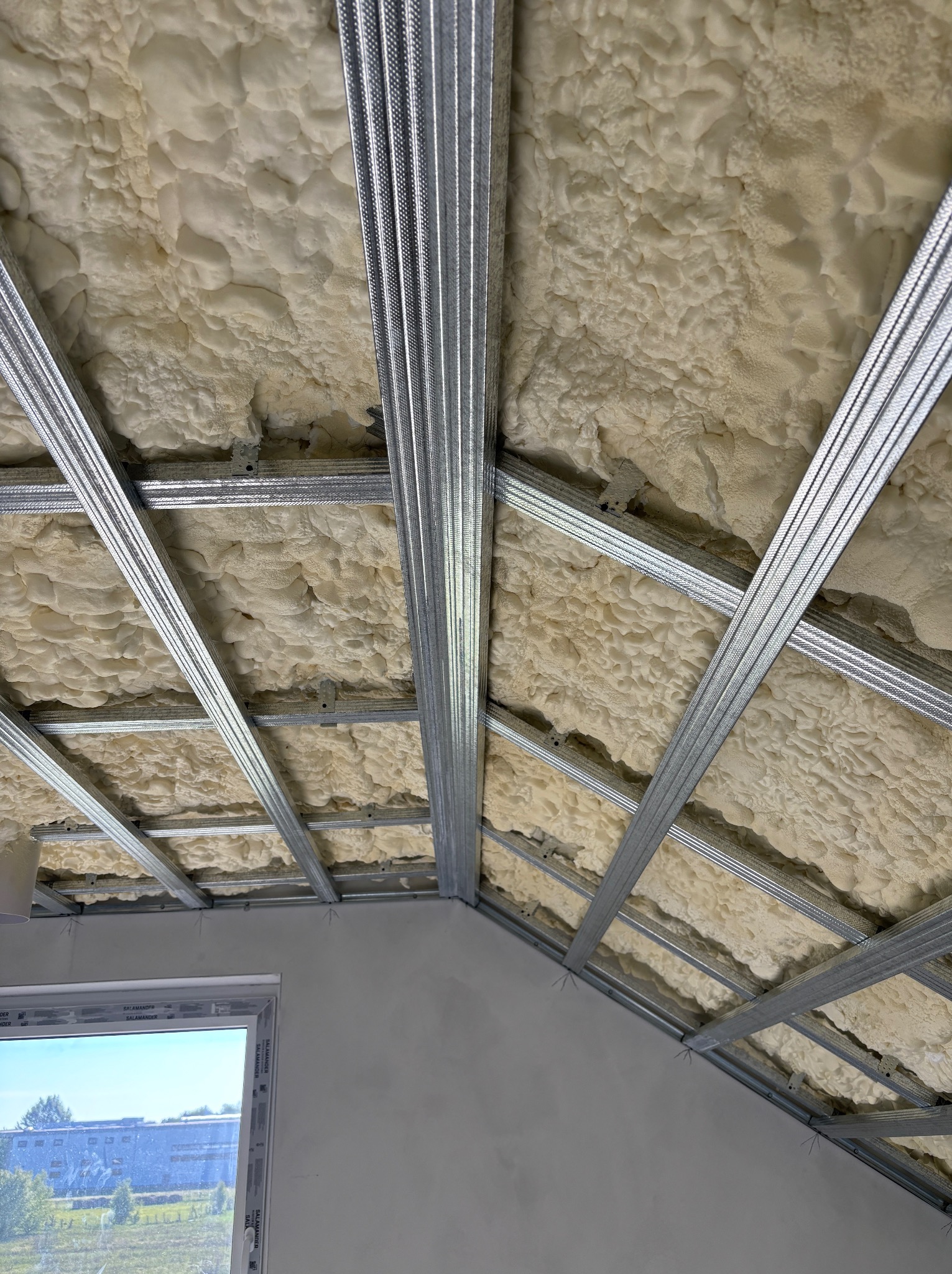 Sufit podwieszany z metalową konstrukcją i pianką izolacyjną, widok z dołu, okno w lewym dolnym rogu. Detal montażu profili i wypełnienia izolacją.