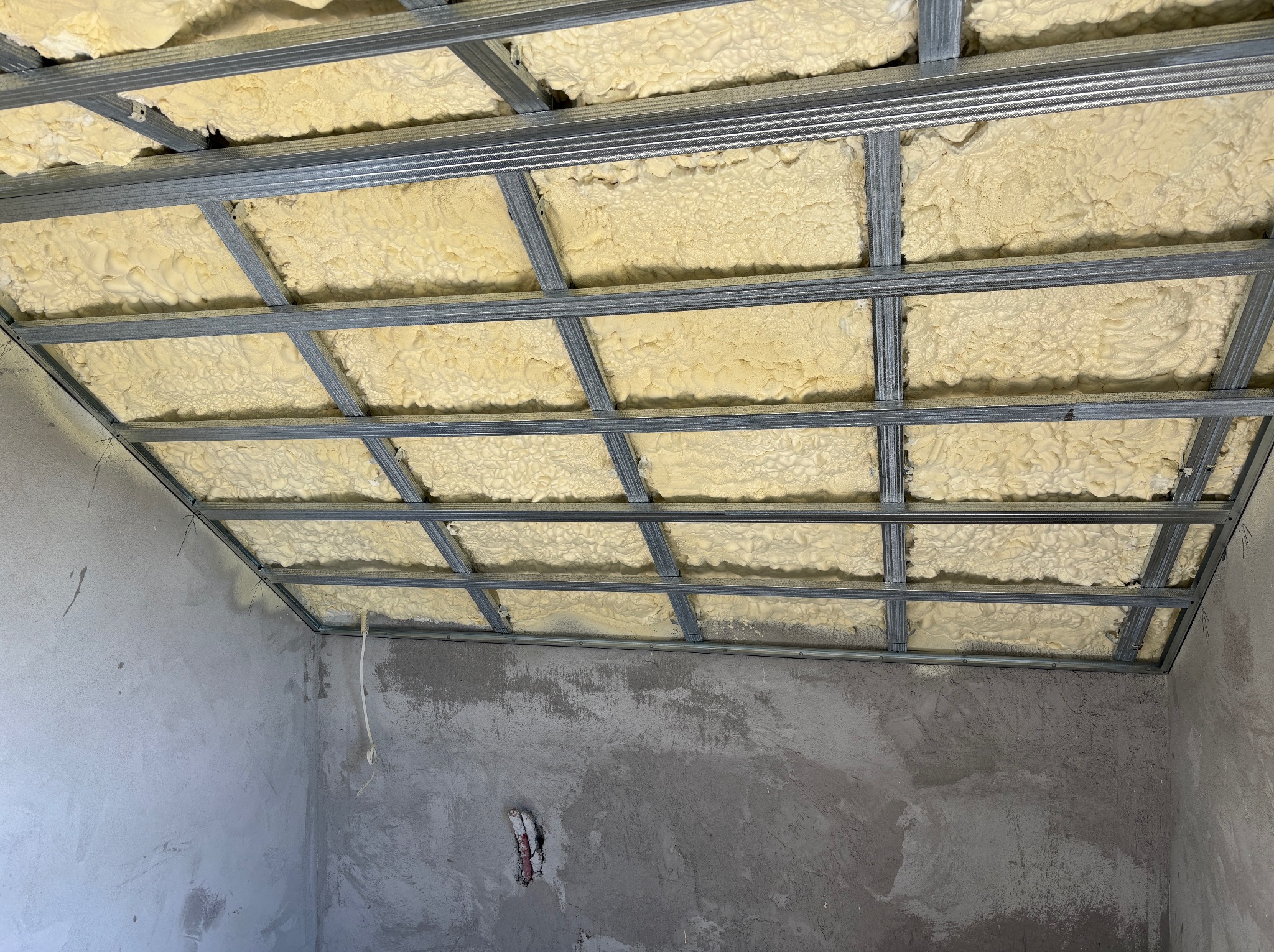 Metalowa konstrukcja sufitu podwieszanego z wypełnieniem pianką izolacyjną na tle surowych ścian. Widoczne elementy instalacji elektrycznej.