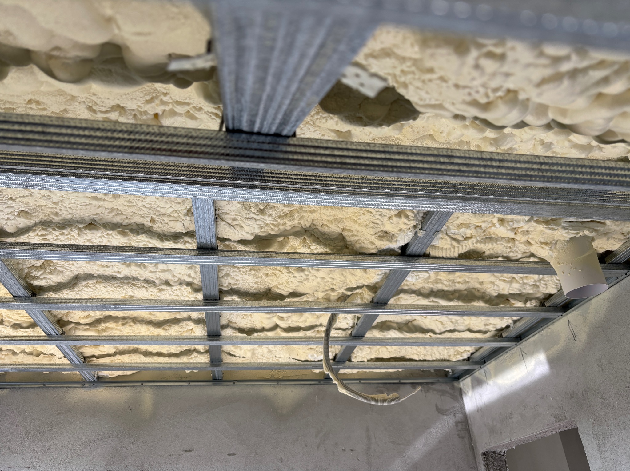 Sufit podwieszany z izolacją piankową, widok z dołu. Metalowa konstrukcja kratowa z widocznymi elementami instalacji, surowy stan wykończenia ścian.