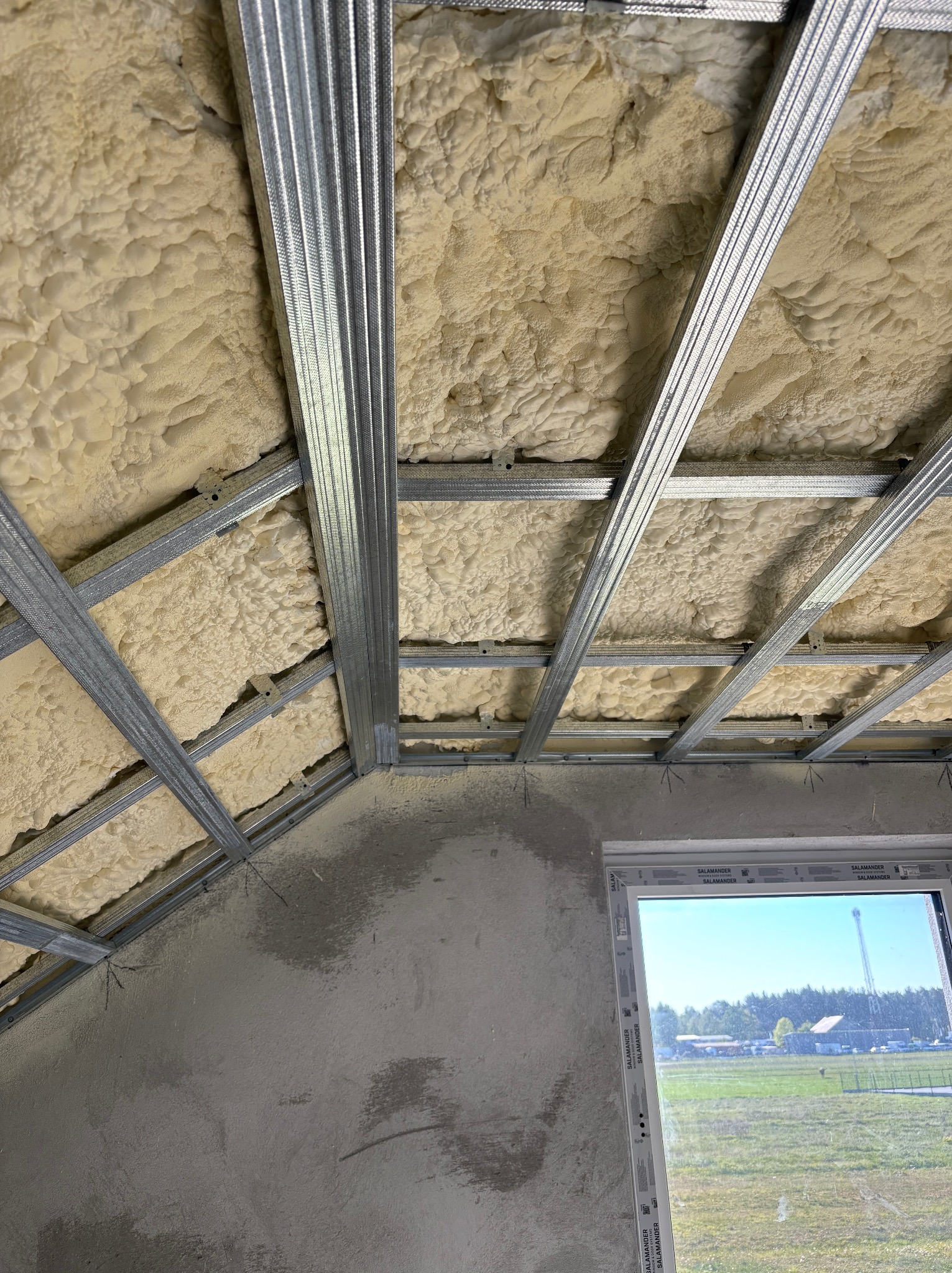 Izolacja poddasza pianą PUR z metalowym stelażem. Widok z dołu na skośny sufit z oknem w tle. Szara ściana w stanie surowym.