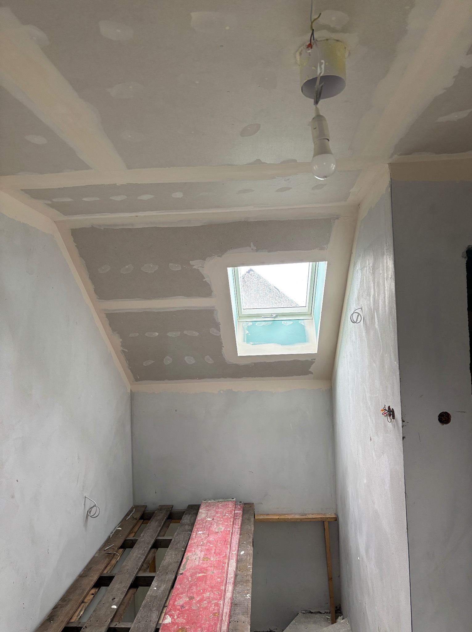 Poddasze ze skośnym sufitem w trakcie prac wykończeniowych. Widoczne płyty gipsowe, szpachlowane łączenia i okno dachowe. Podłoga zabezpieczona paletami.