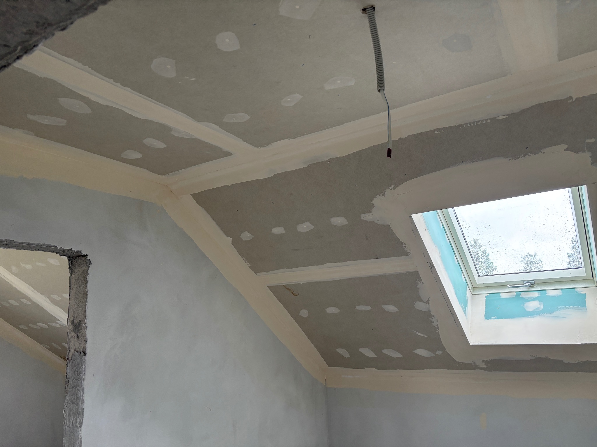 Poddasze w trakcie wykańczania: sufit z płyt gipsowych, szpachlowane łączenia, okno dachowe. Widoczny przewód elektryczny. Ściany częściowo wykończone.