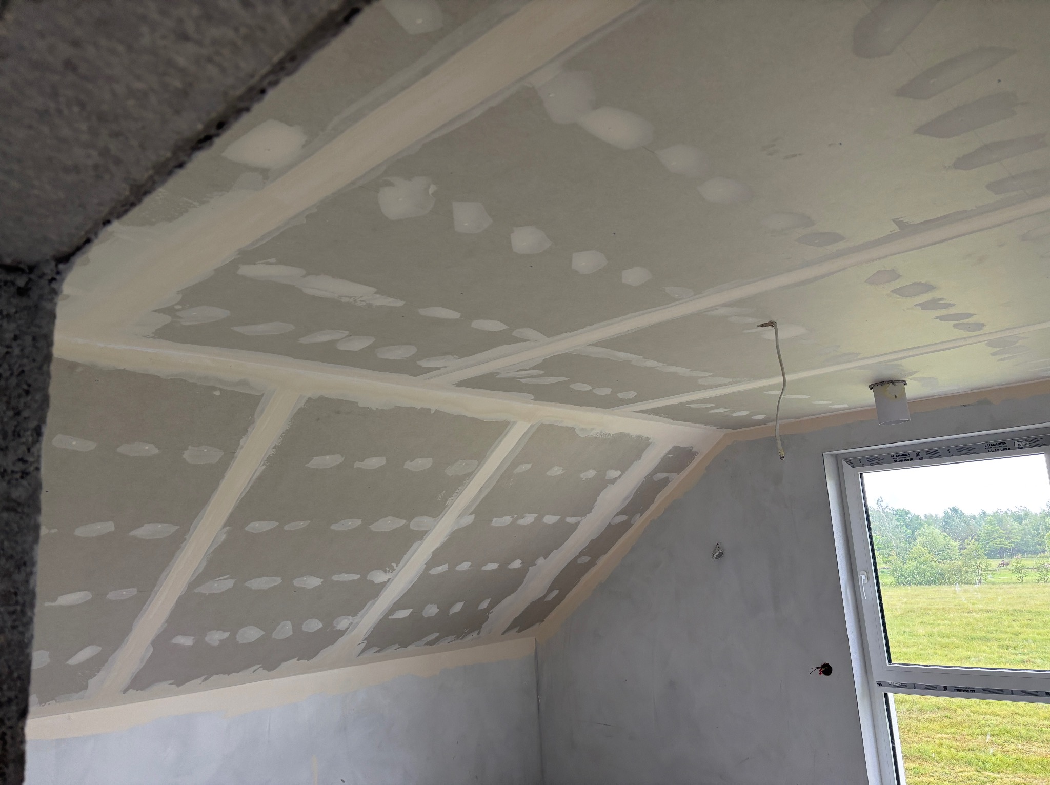 Skośny sufit poddasza po montażu płyt gipsowo-kartonowych i szpachlowaniu łączeń, widok z dołu, fragment okna i zielony krajobraz w tle.