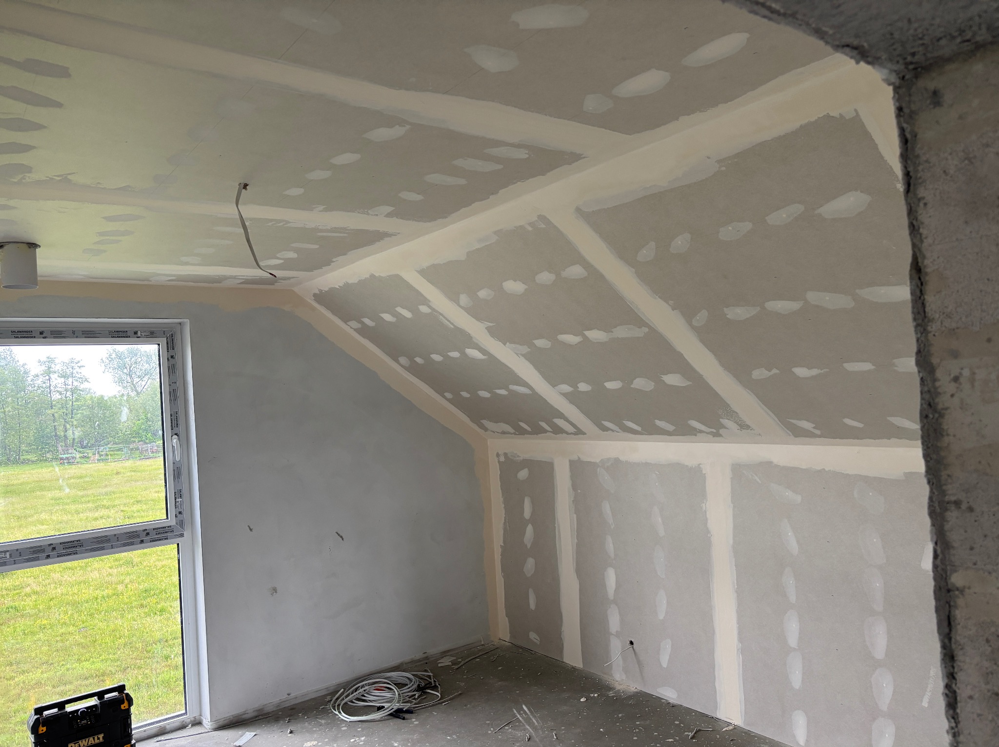 Montaż płyt gipsowo-kartonowych na poddaszu ze skosami, widoczne łączenia płyt i szpachlowanie. Okno z widokiem na zieleń, narzędzia DeWalt na podłodze.