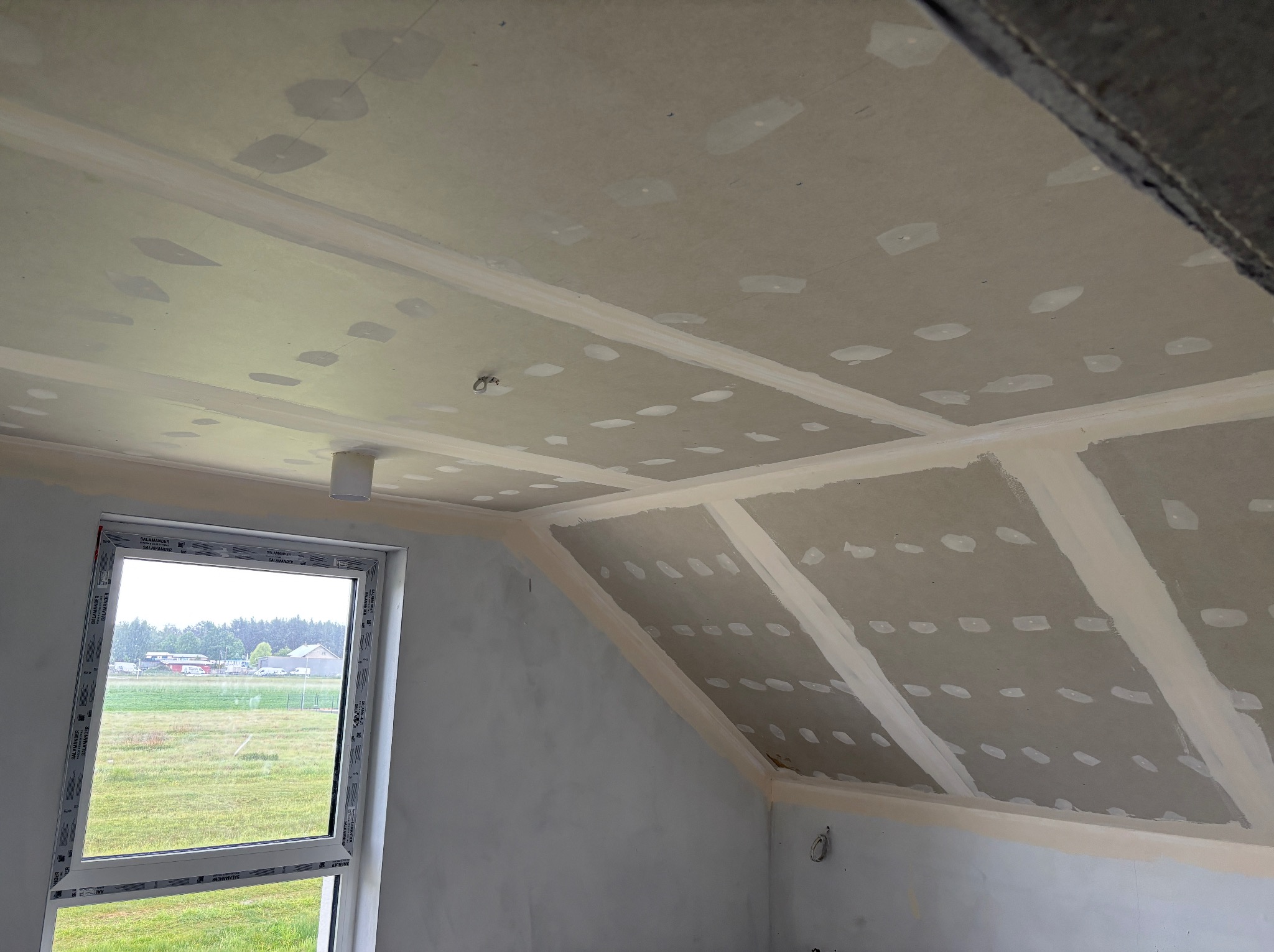 Montaż płyt gipsowych na poddaszu w Jasienicy. Widoczne łączenia płyt i szpachlowane wkręty. Okno z widokiem na zewnątrz.