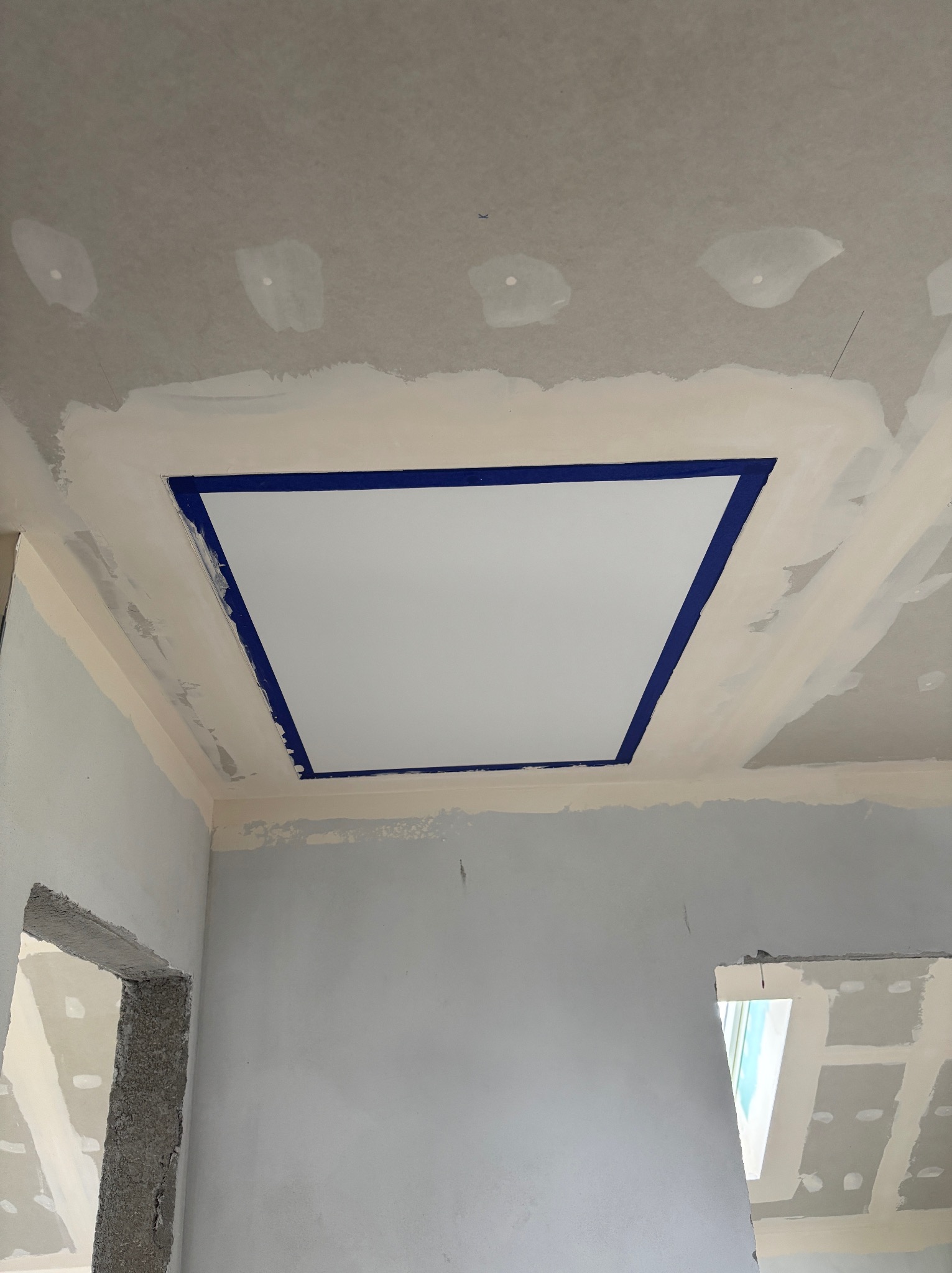 Nierówna powierzchnia sufitu z płyt kartonowo-gipsowych w trakcie szpachlowania, widoczne niebieskie taśmy malarskie wokół kwadratowej wnęki.