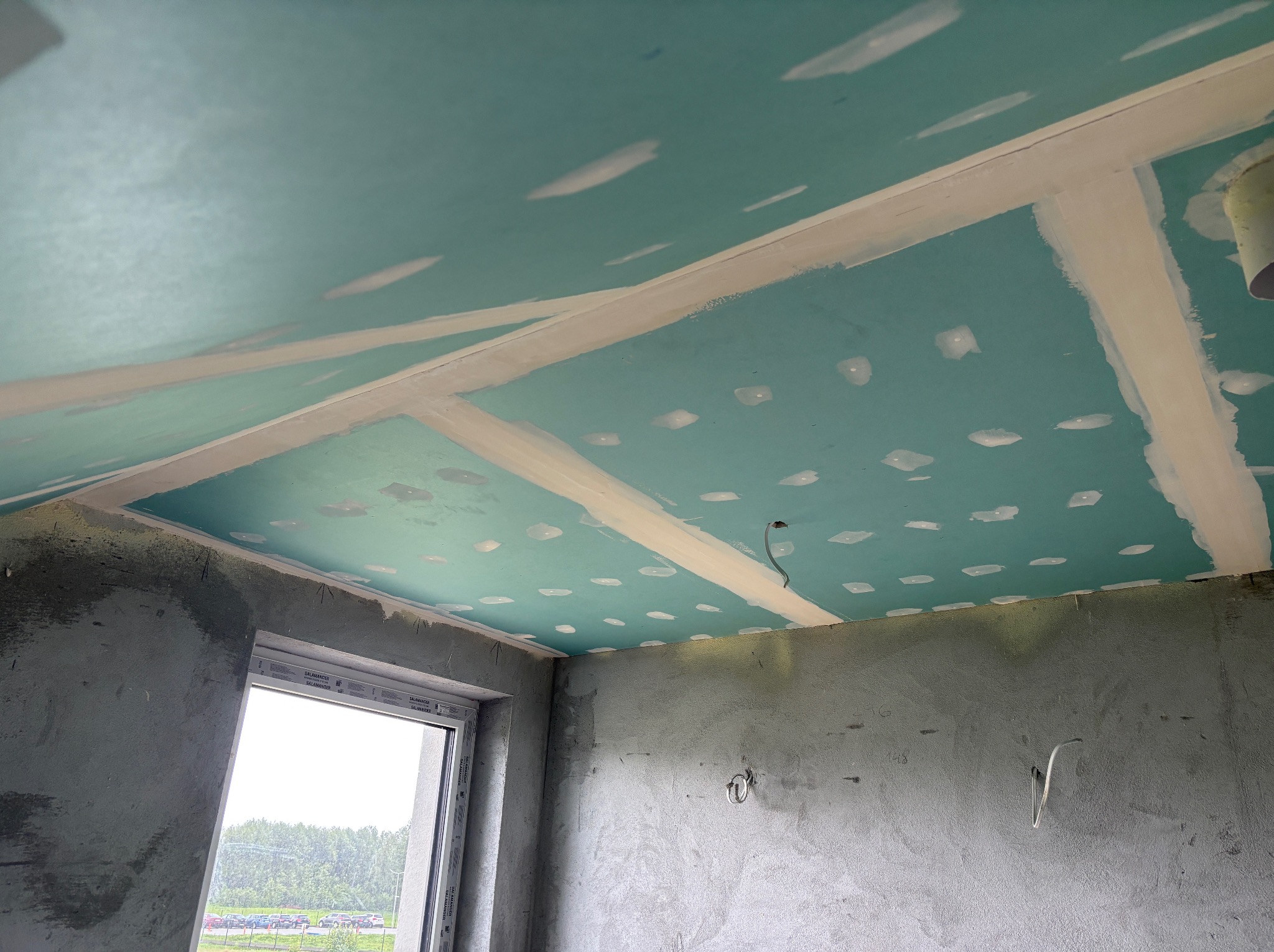 Niebieski sufit z płyt kartonowo-gipsowych, z widocznymi łączeniami i szpachlowanymi wkrętami, surowe ściany i okno z widokiem na zieleń.