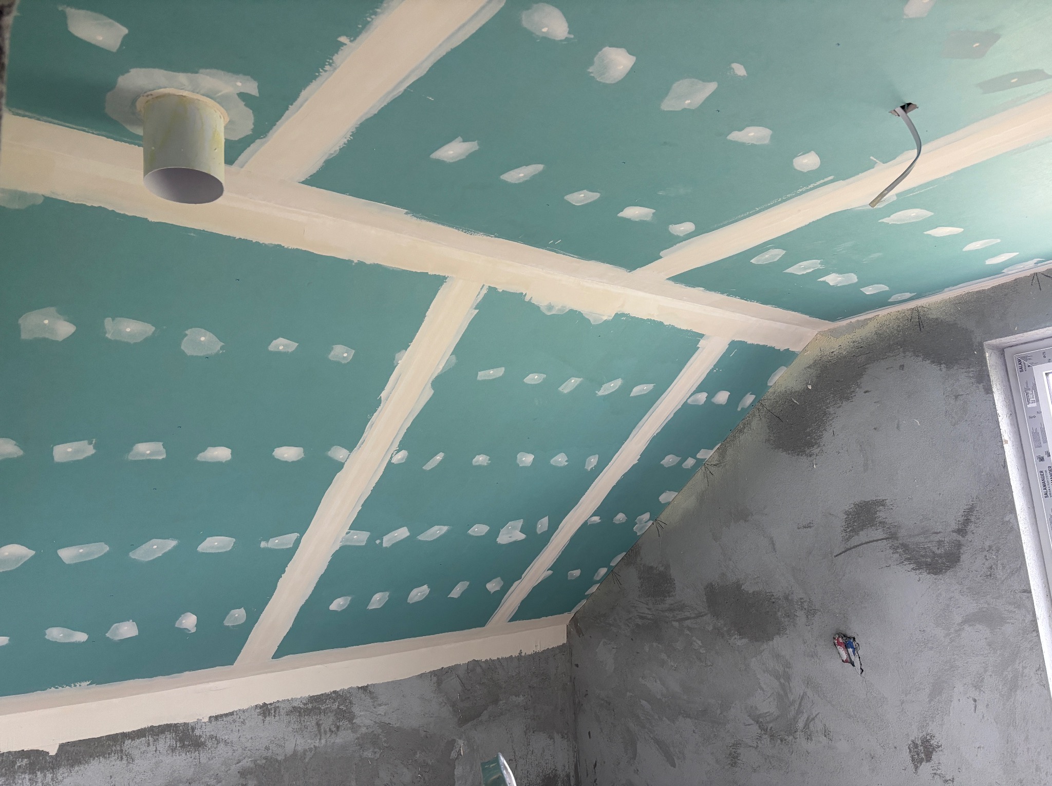 Płyty gipsowo-kartonowe na skośnym suficie poddasza, z widocznymi łączeniami i miejscami po wkrętach, przygotowane do szpachlowania. Ściana surowa, okno w rogu.