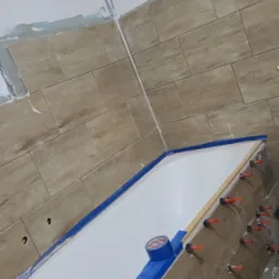 Układanie płytek imitujących drewno wokół wanny, z użyciem poziomowania i taśmy maskującej. Widoczne narzędzia glazurnicze i fragment ściany z częściowo położonymi płytkami.