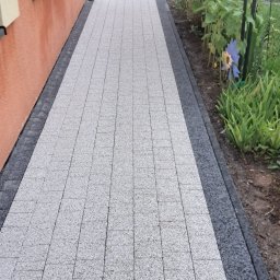 Gospodarstwo Ogrodnicze Krzysztof Dudek - Jasnoszara kostka brukowa ułożona wzdłuż pomarańczowej ściany budynku, obramowana ciemniejszą kostką. W tle widoczna zieleń ogrodu i fragment rynny.